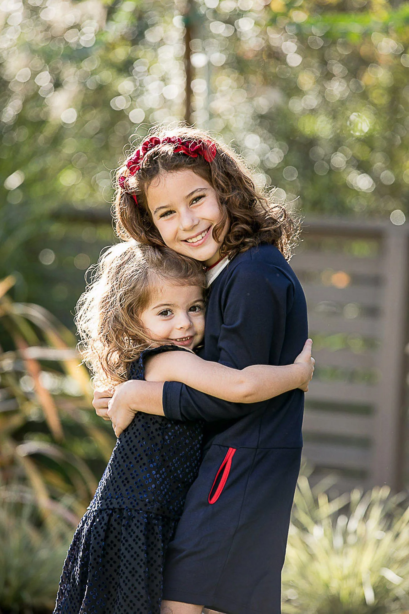 Two girls hugging outdoors with sunlight creating bokeh background, one with a red headband and curly hair, the other with straight hair and a black dress.
