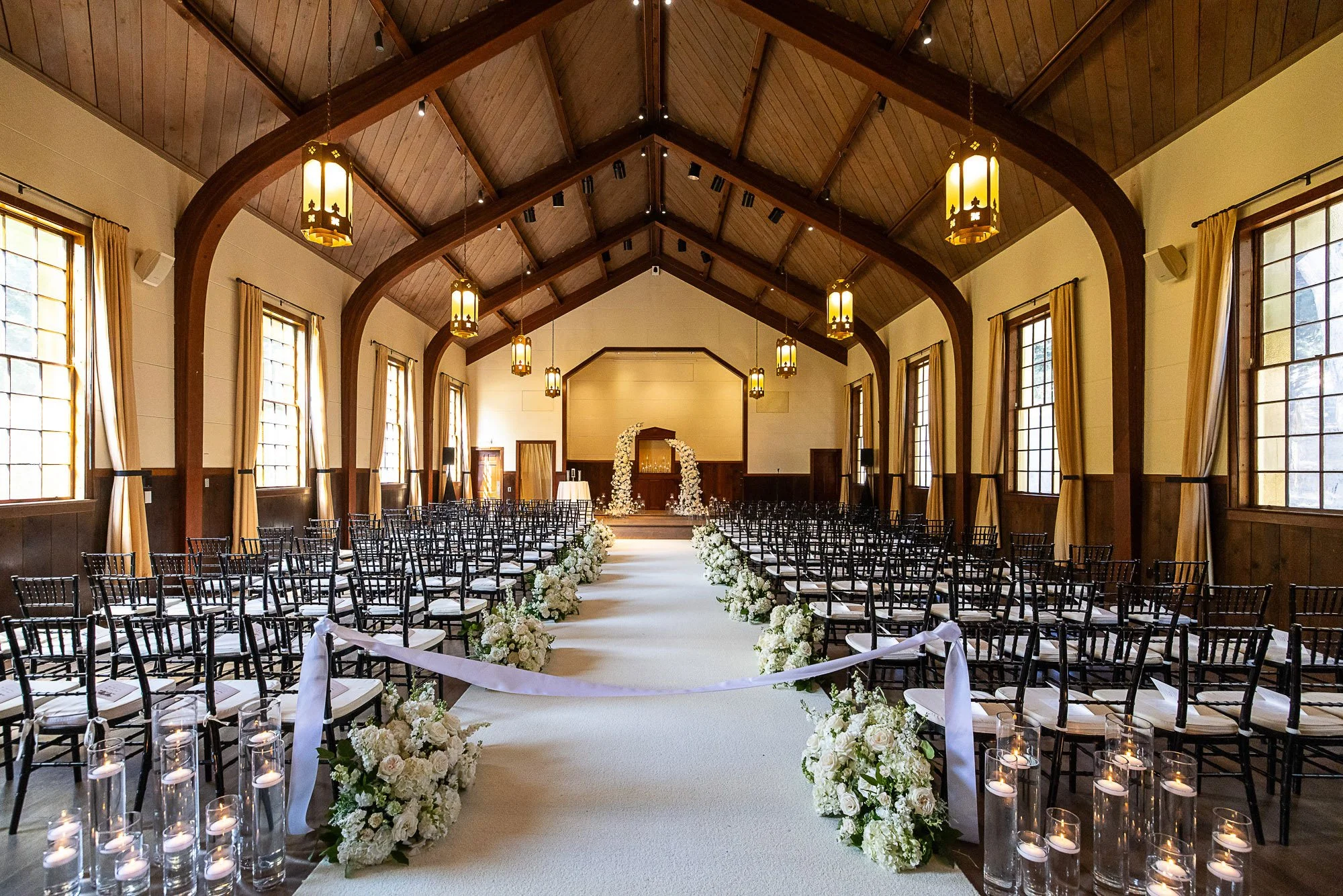 An indoor wedding ceremony setup with rows of black chairs, an aisle lined with white floral arrangements, and a decorated arch at the front. The room has wooden ceilings with hanging lanterns and large windows with curtains.
