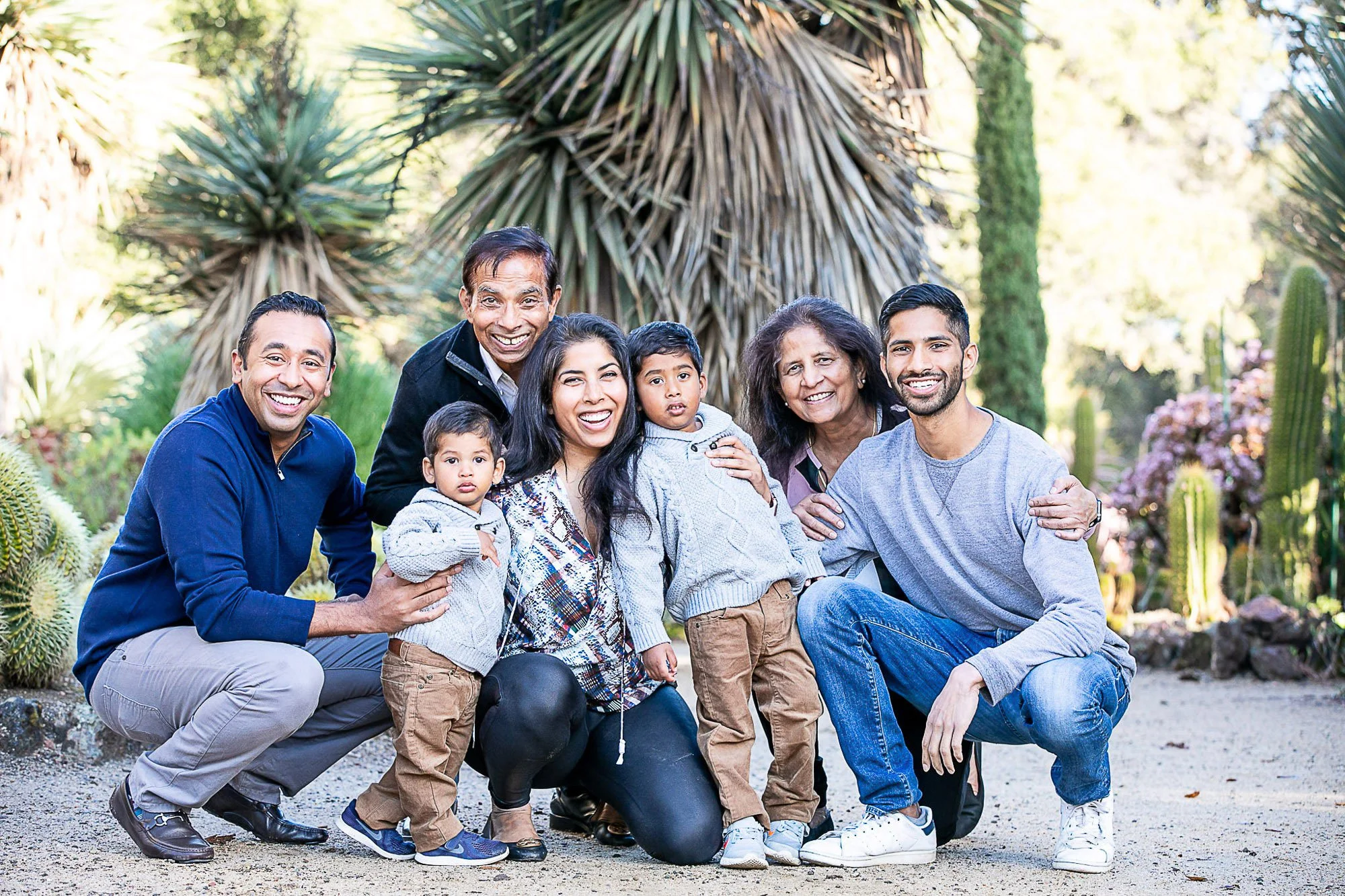 A diverse family group of seven people, including children and adults, smiling and posing together outdoors in a botanical garden with various desert plants and cacti in the background.