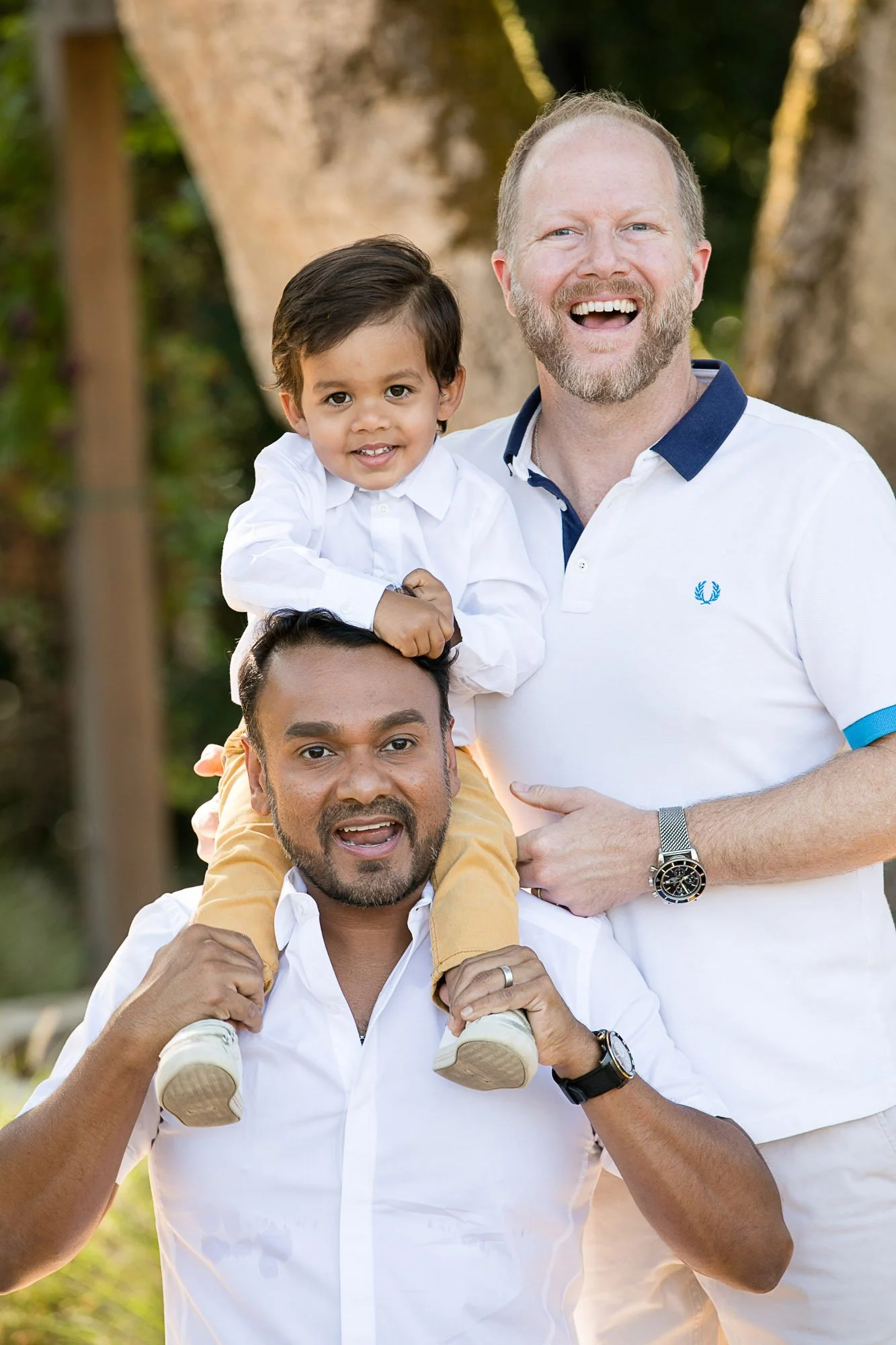 A happy man with a beard on his face being carried on shoulders by another man, with a young girl smiling and sitting on his shoulders, in a park with trees in the background.