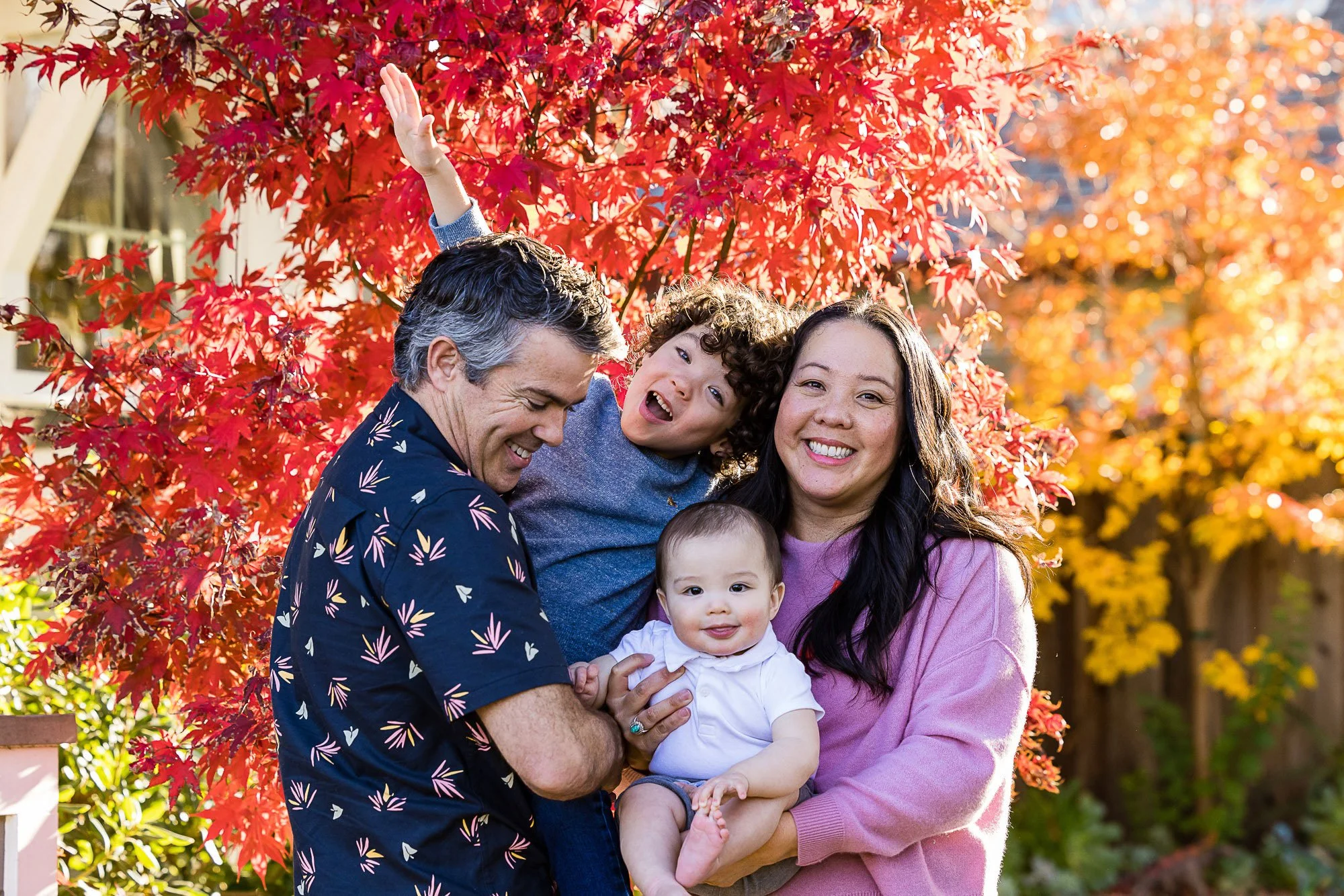 A family of five smiling together outdoors in front of vibrant red and orange autumn leaves, with a man, woman, two children, and a baby.