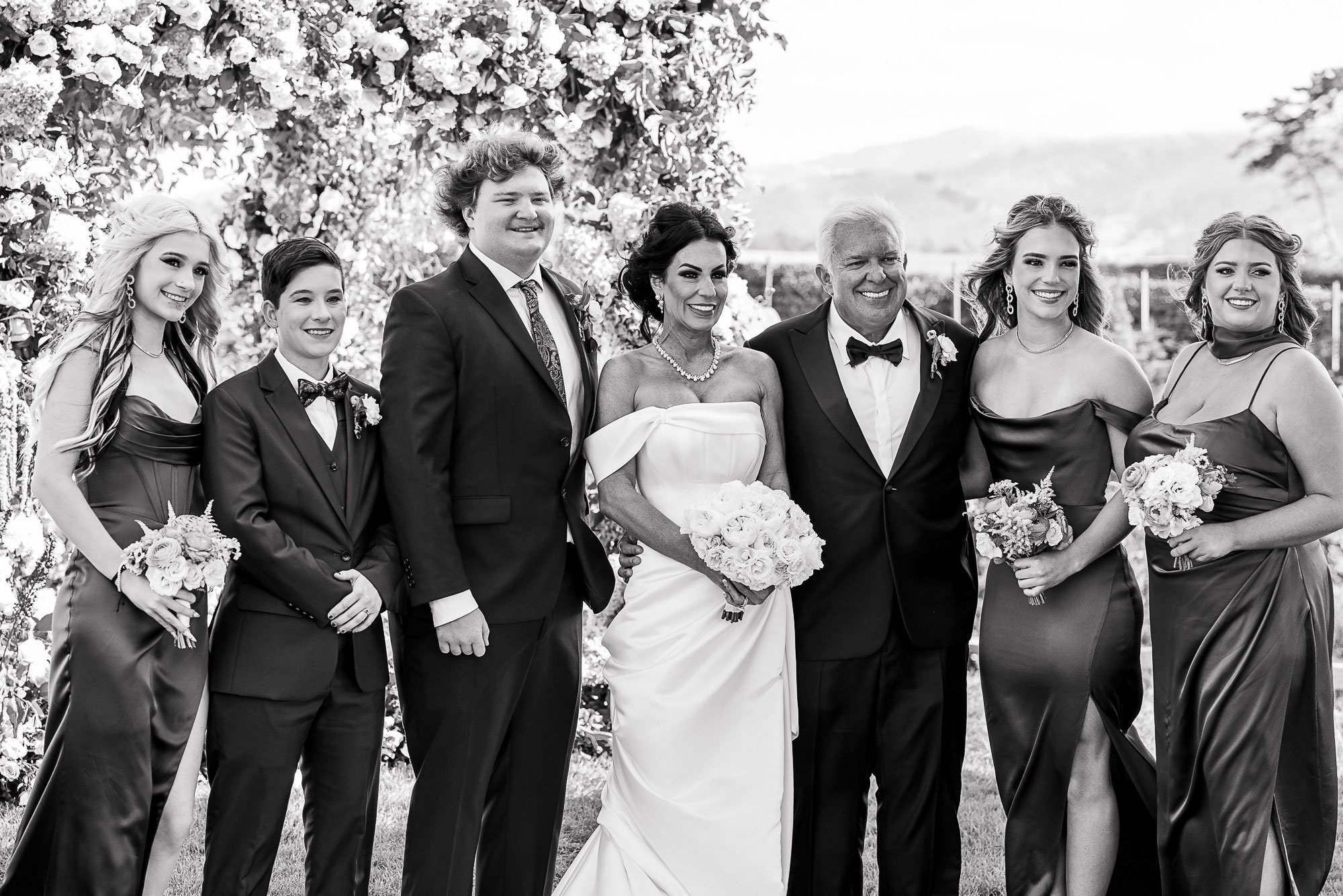 Group photo at a wedding with seven people, including bride and groom, standing outdoors with floral arch backdrop at the Beach and Tennis Club in Pebble Beach, CA.