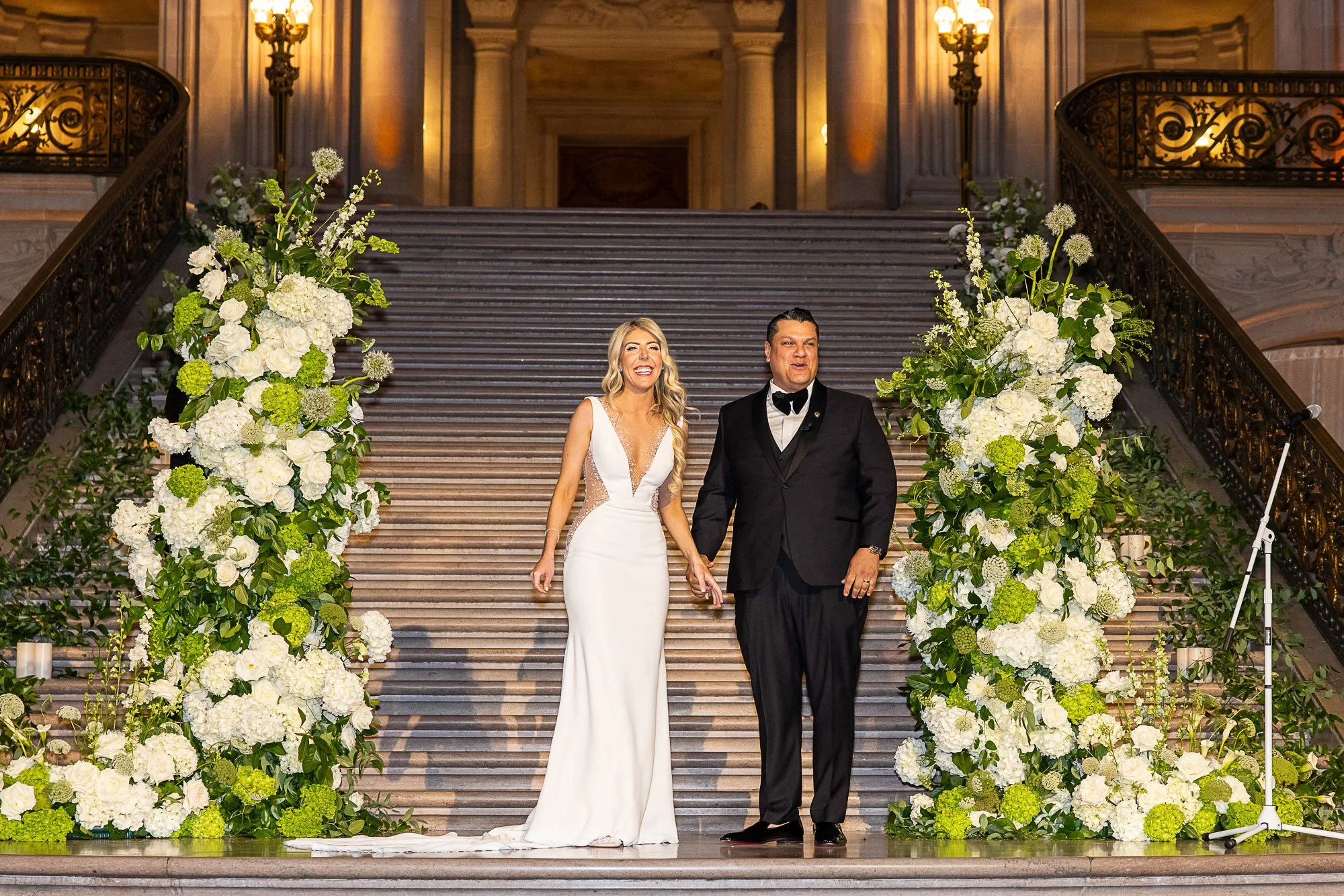 Bride and groom descending the grand staircase at San Francisco City Hall between white floral arrangements