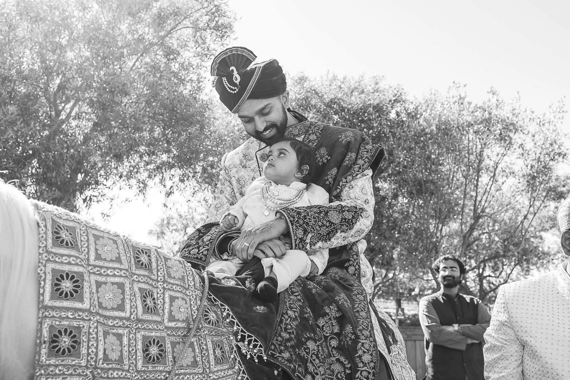A man in traditional Indian attire holding a young girl on his lap during a cultural celebration outdoors, with two other men in the background and trees.