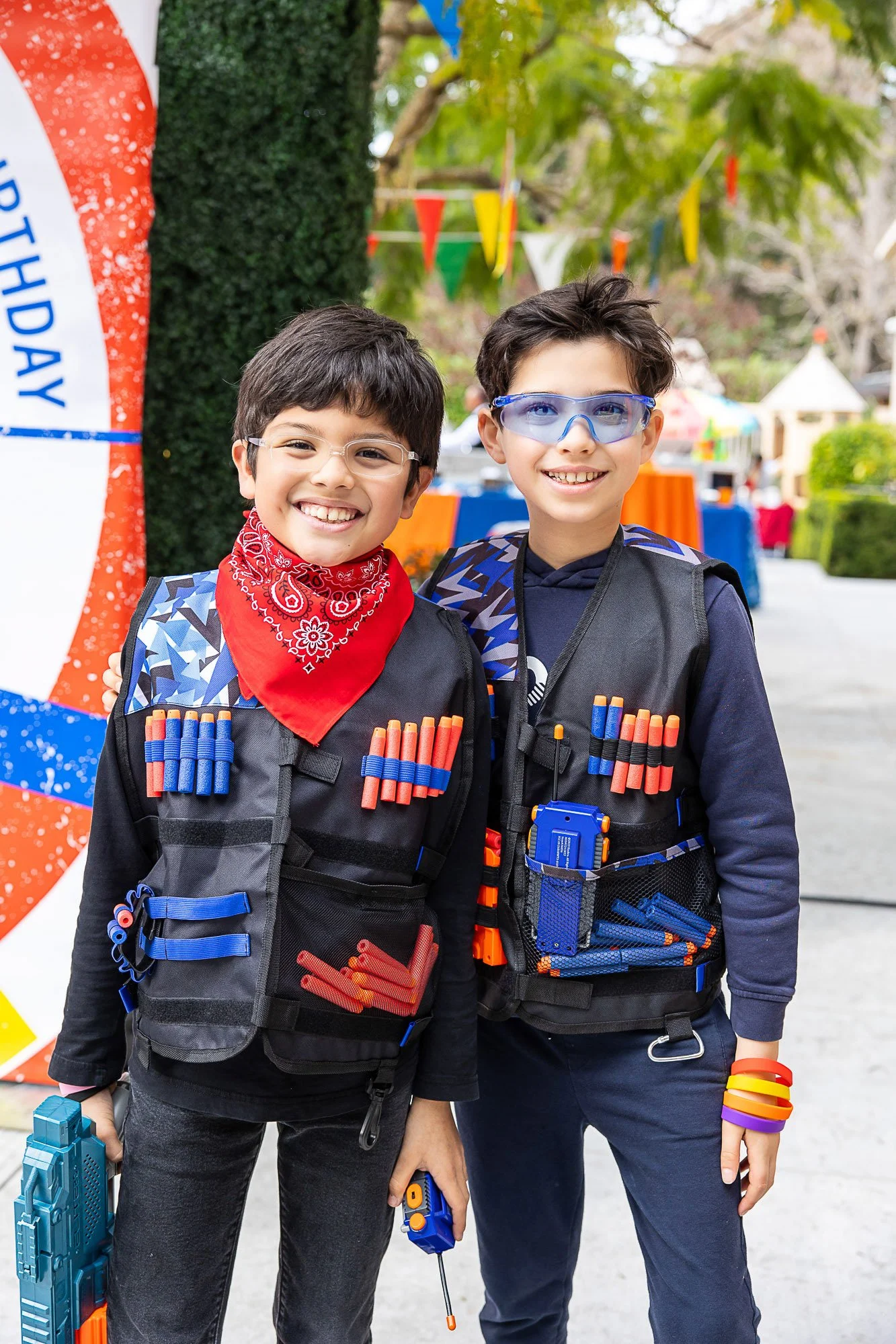 Two boys smiling at a theme park, wearing black vests with foam darts, glasses, and holding foam dart guns. Colorful banners and park structures are visible in the background.