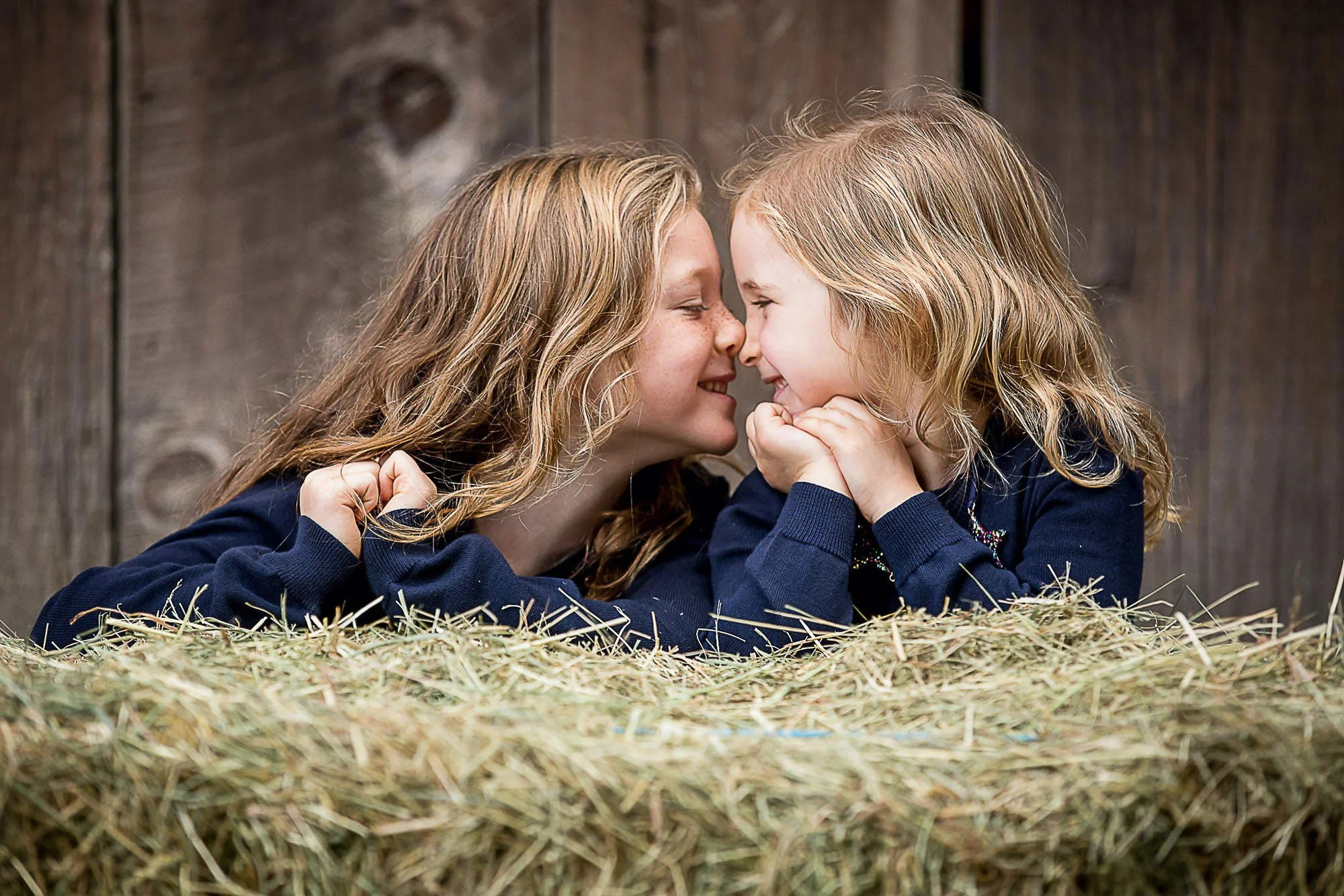 Two young girls with blonde hair and blue eyes lying on straw, facing each other with foreheads touching, smiling and holding each other's faces in a rustic setting.