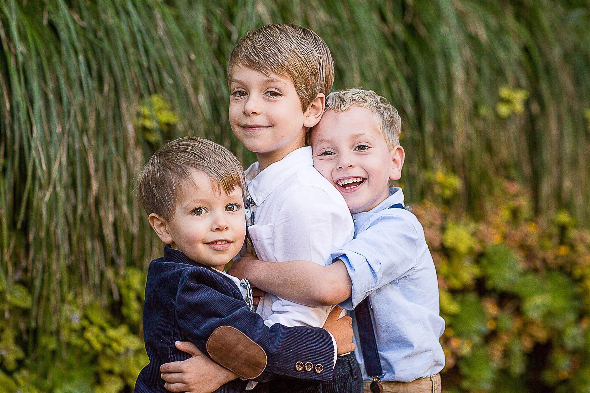 Three young boys hugging outdoors in front of tall grass and trees, all smiling at the camera.