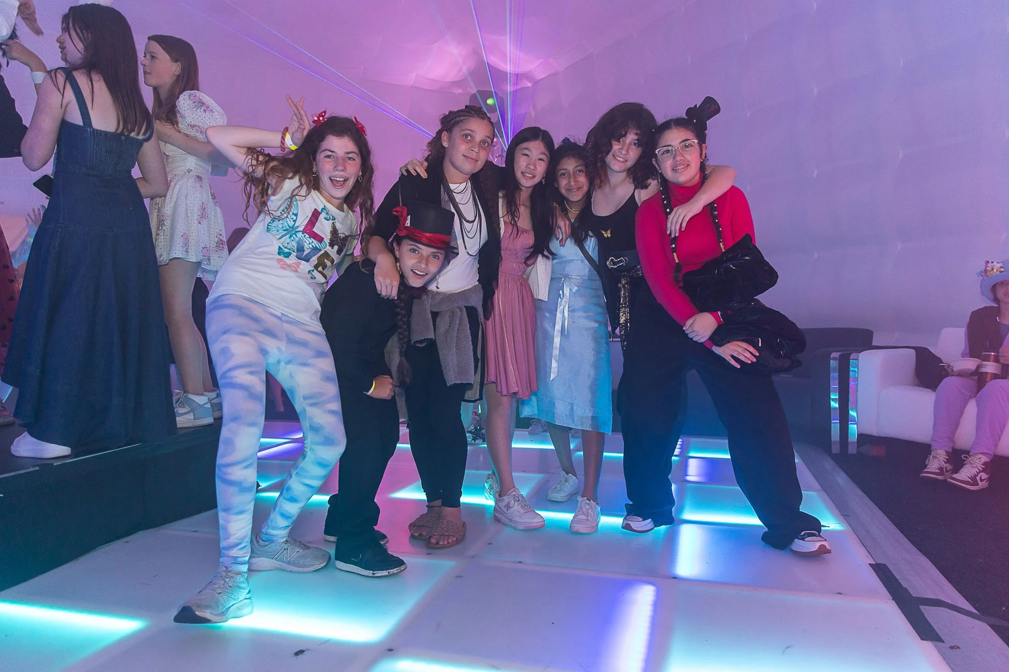 Group of young girls at a party, smiling and posing on a lit platform with colorful lighting and lasers.