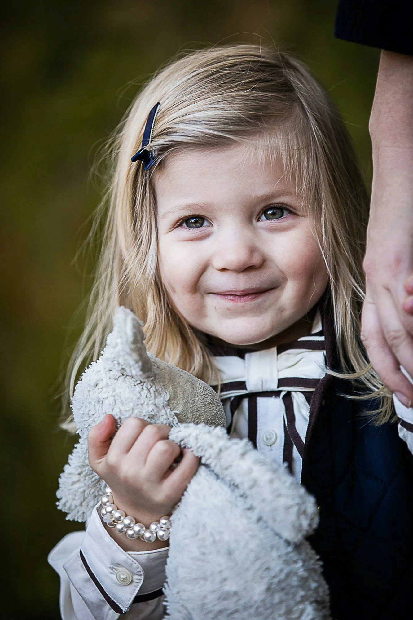A young girl with blonde hair held back with a navy hair clip, smiling and looking up, wearing a white shirt with black trim, a pearl bracelet, and holding a small, fluffy beige stuffed animal. An adult, partially visible, is holding her hand.
