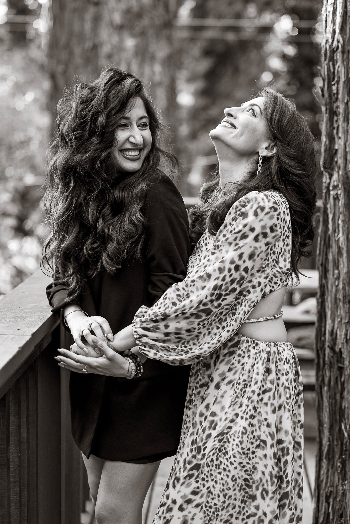 Two women smiling and laughing outdoors near a wooden railing and a tree, one woman wearing a leopard print dress and the other wearing a dark jacket, black and white photo.