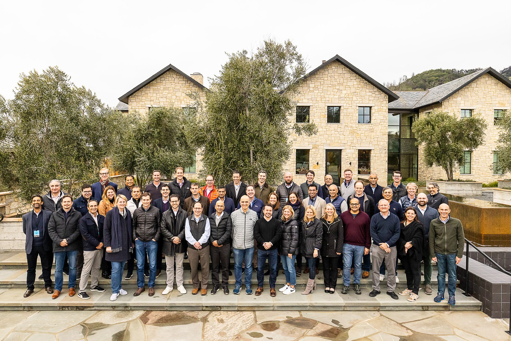 A large group of people standing together outdoors in front of a stone building with trees and a small water feature in the background at the Napa Valley Four Seasons Hotel.