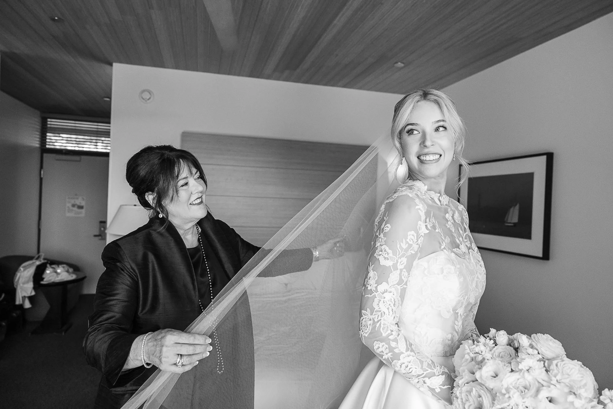 A bride with blonde hair wearing a lace wedding dress, holding a bouquet of white roses, smiling as an older woman helps her with a veil in a modern room with wood-paneled ceiling at Cavallo Point in Sausalito, CA..