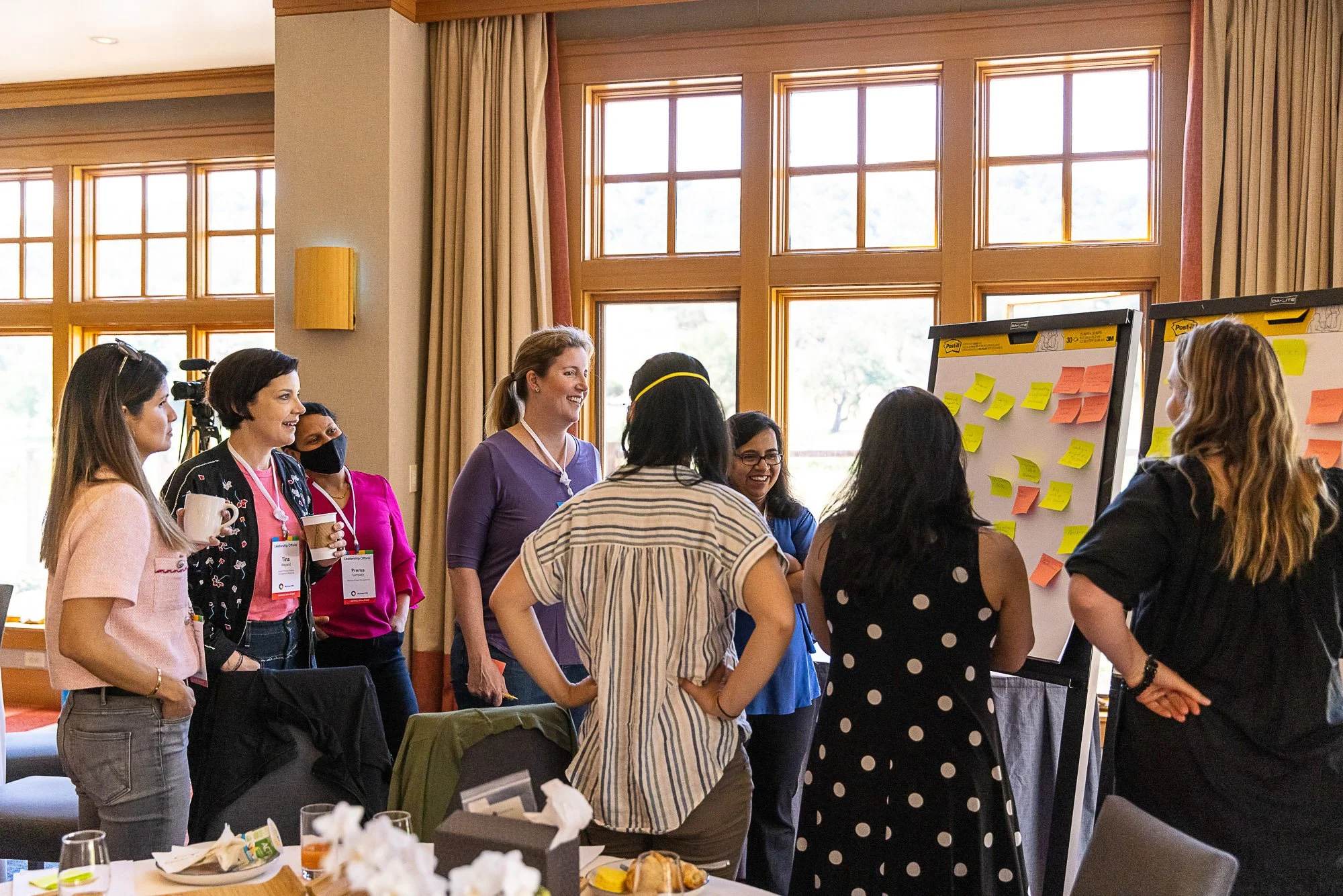 Group of women gathered around a whiteboard with sticky notes, engaged in discussion at a conference or workshop in a well-lit room with large windows.