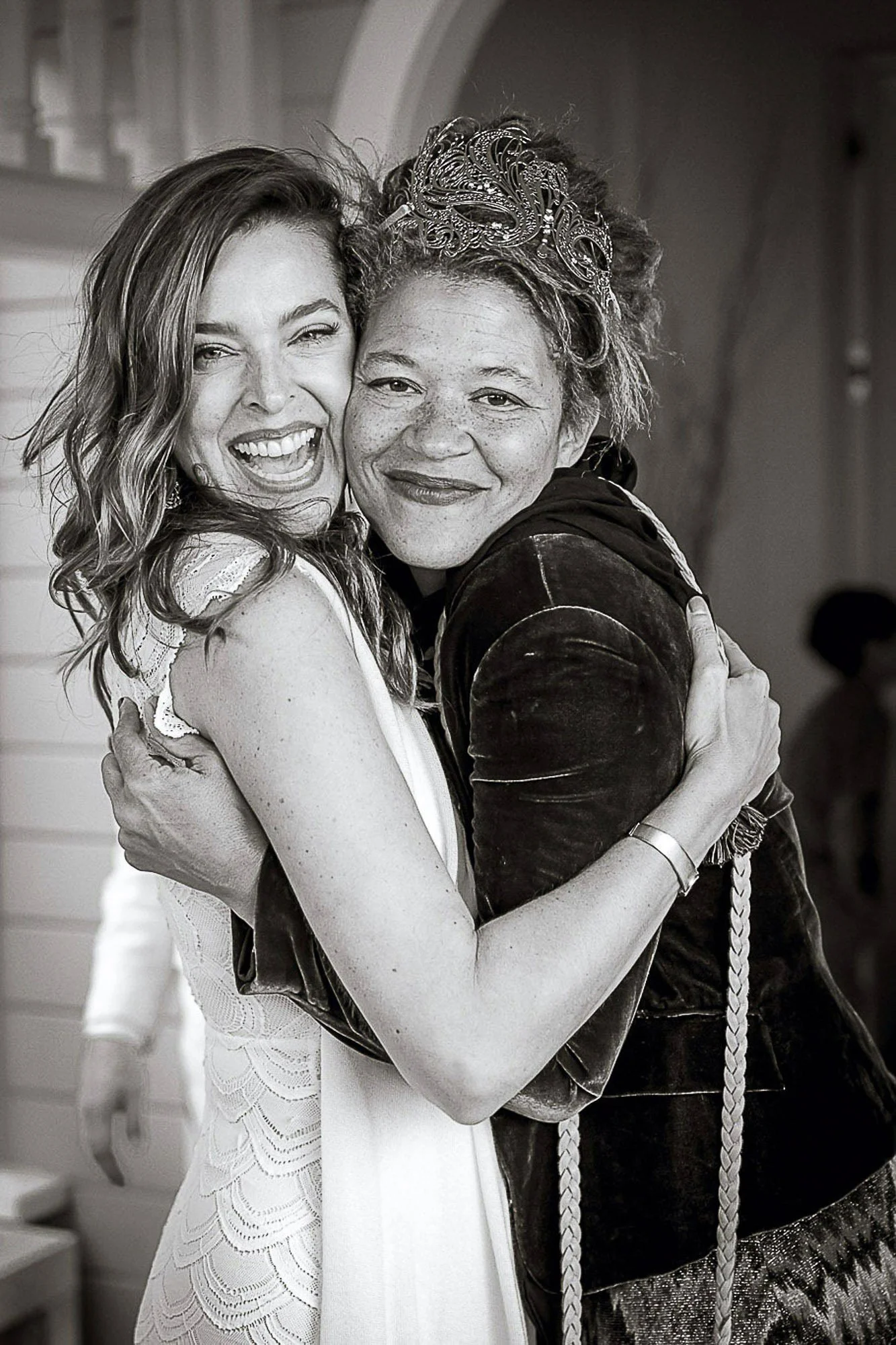 Two women hugging and smiling, one with light hair and the other with curly hair and a decorative headband, in a black-and-white photo.