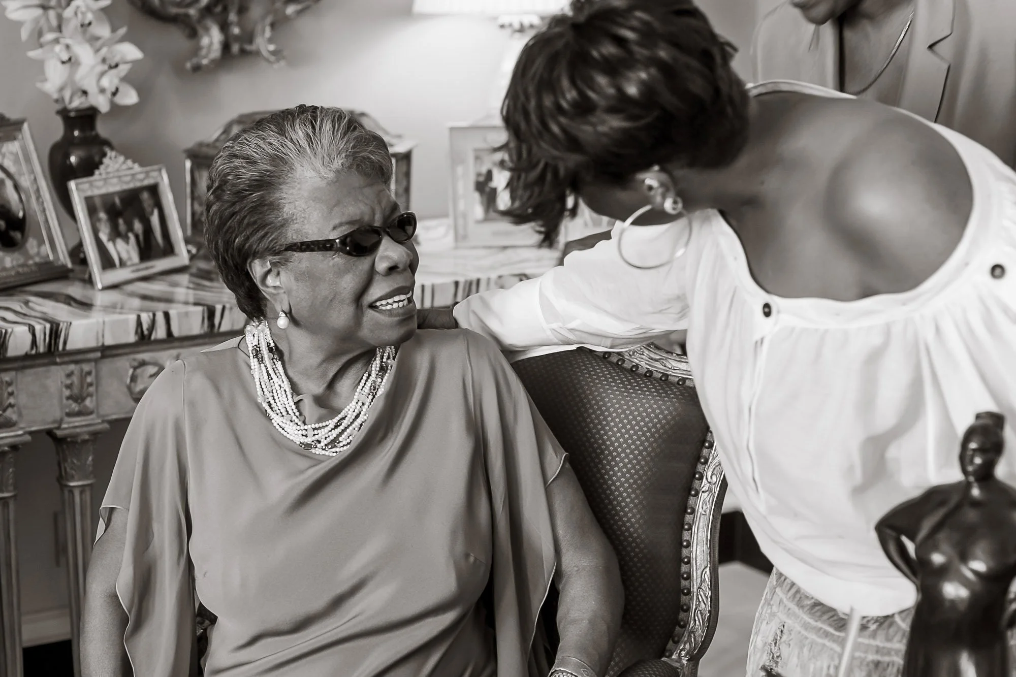 Maya Angelou smiling and interacting in a living room. One woman, older with short hair and glasses, wearing a necklace and earrings, is seated. The other woman, younger with wavy hair, wearing a white blouse, is leaning toward her. 