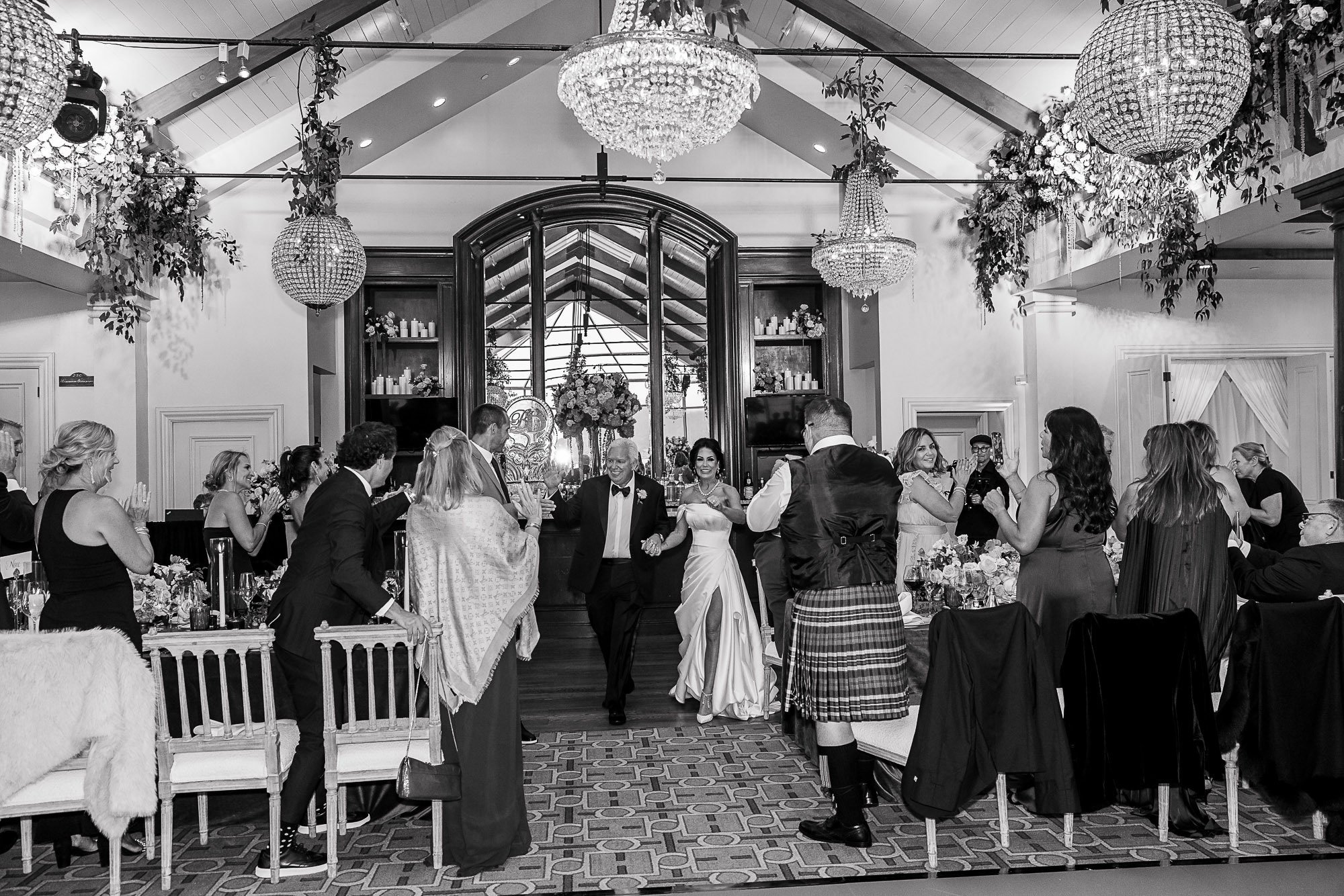 A wedding reception at the Beach and Tennis Club in Pebble Beach, CA with guests dancing and celebrating. Elegant decor with floral arrangements, chandeliers, and a large mirror behind the bar area.