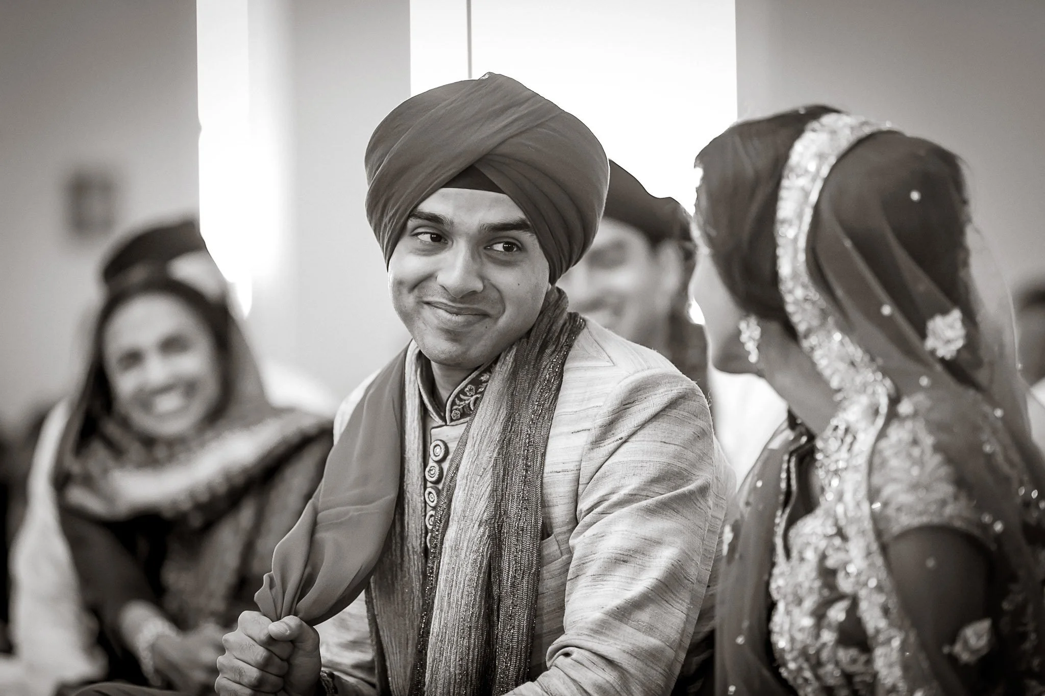 A man wearing traditional Indian attire, including a turban and embroidered clothing, sitting with others dressed in similar festive clothing, likely during a cultural celebration or wedding.