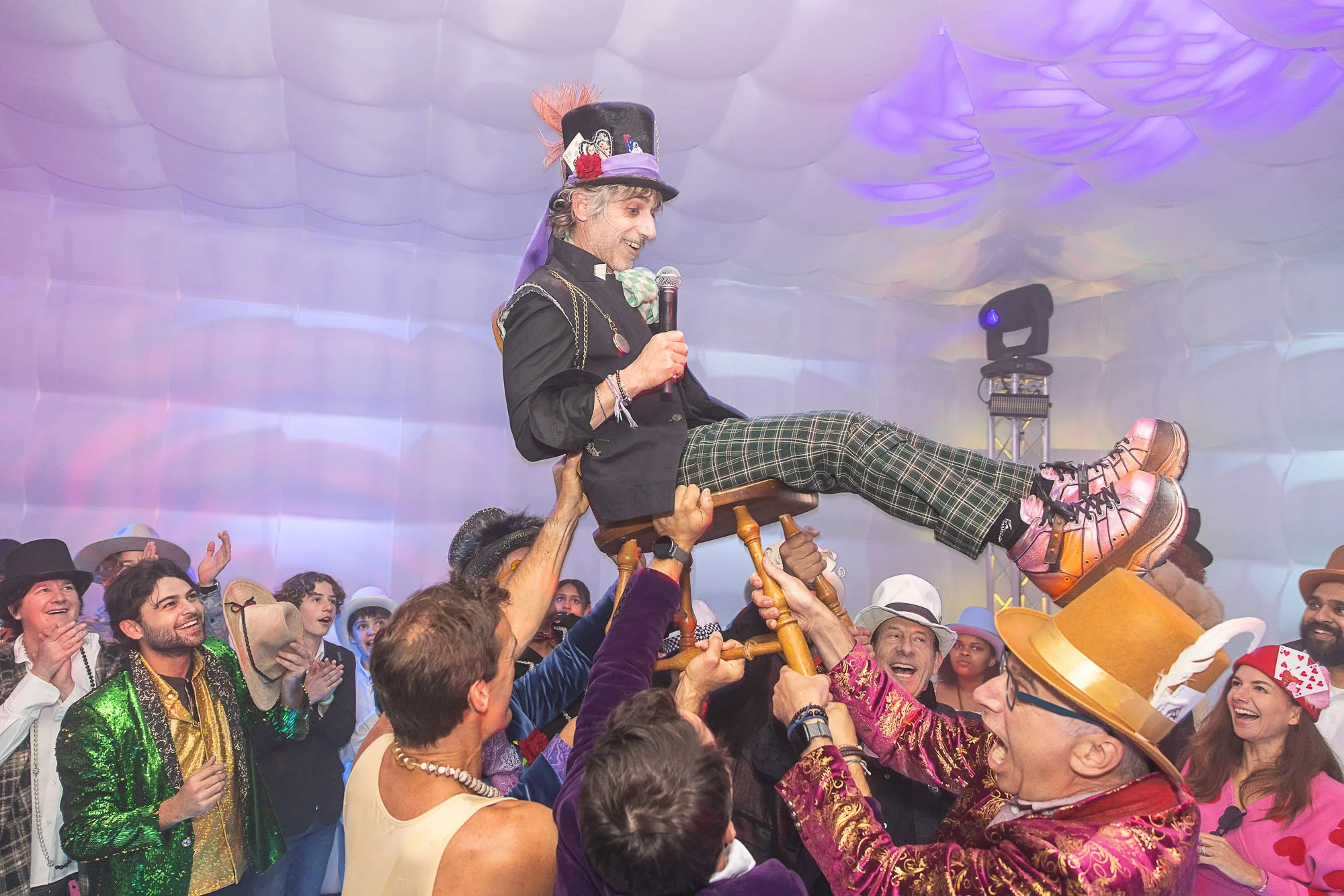 A man dressed in colorful costume, sitting on a chair held up by several people at a lively costume party or celebration, with many smiling and cheering people around at a bat mitzvah.