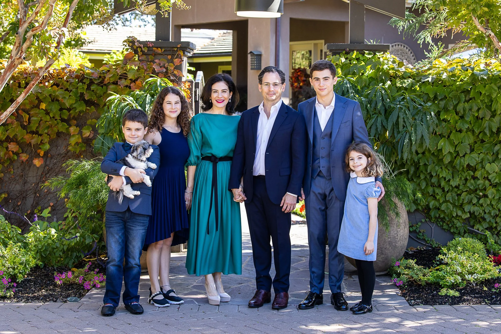 A family of seven standing outdoors in front of a house, holding hands, with trees and greenery around. The group includes three adults and four children, one of whom is holding a puppy. Everyone is dressed nicely and smiling.
