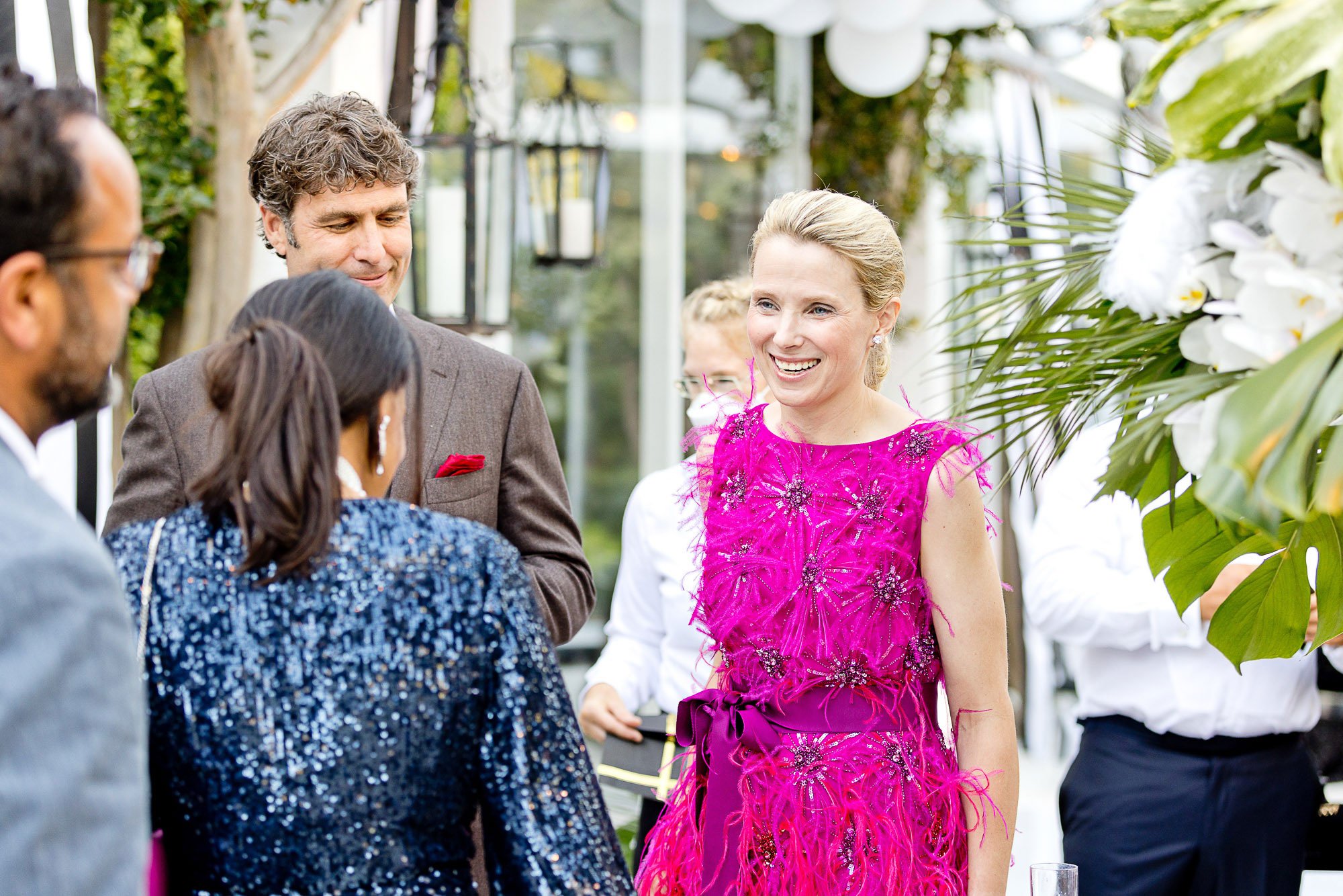 Marissa Mayer in a bright pink dress with intricate embroidery is talking to a group of people at a social gathering outdoors. Men and women are dressed in formal attire, and the setting features greenery and decorative hanging lanterns.