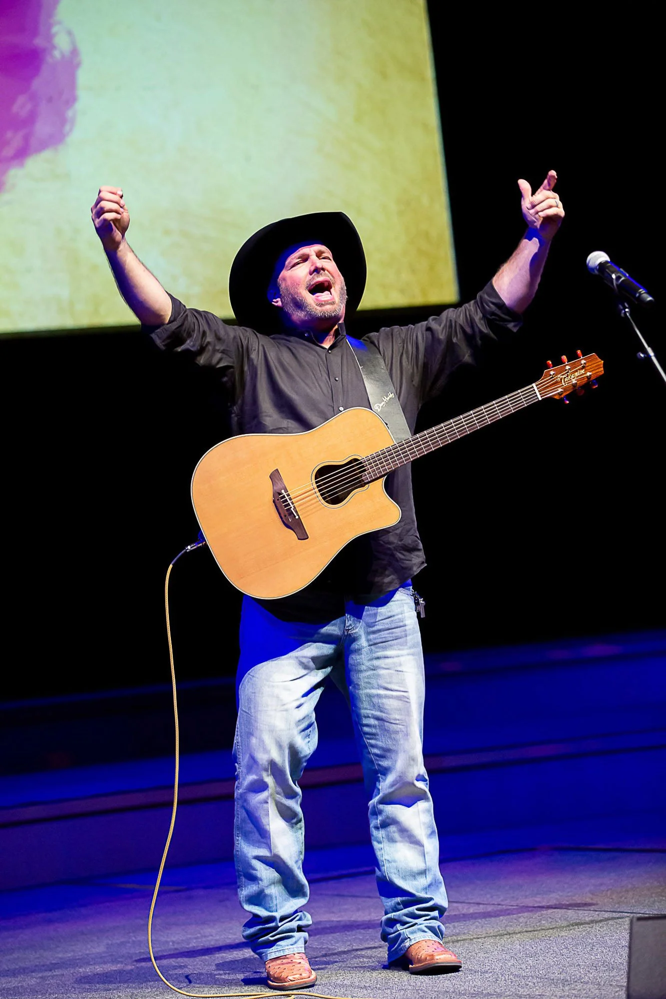 Garth Brooks singing passionately while playing an acoustic guitar on stage, wearing a black cowboy hat, black shirt, light-colored jeans, and cowboy boots.
