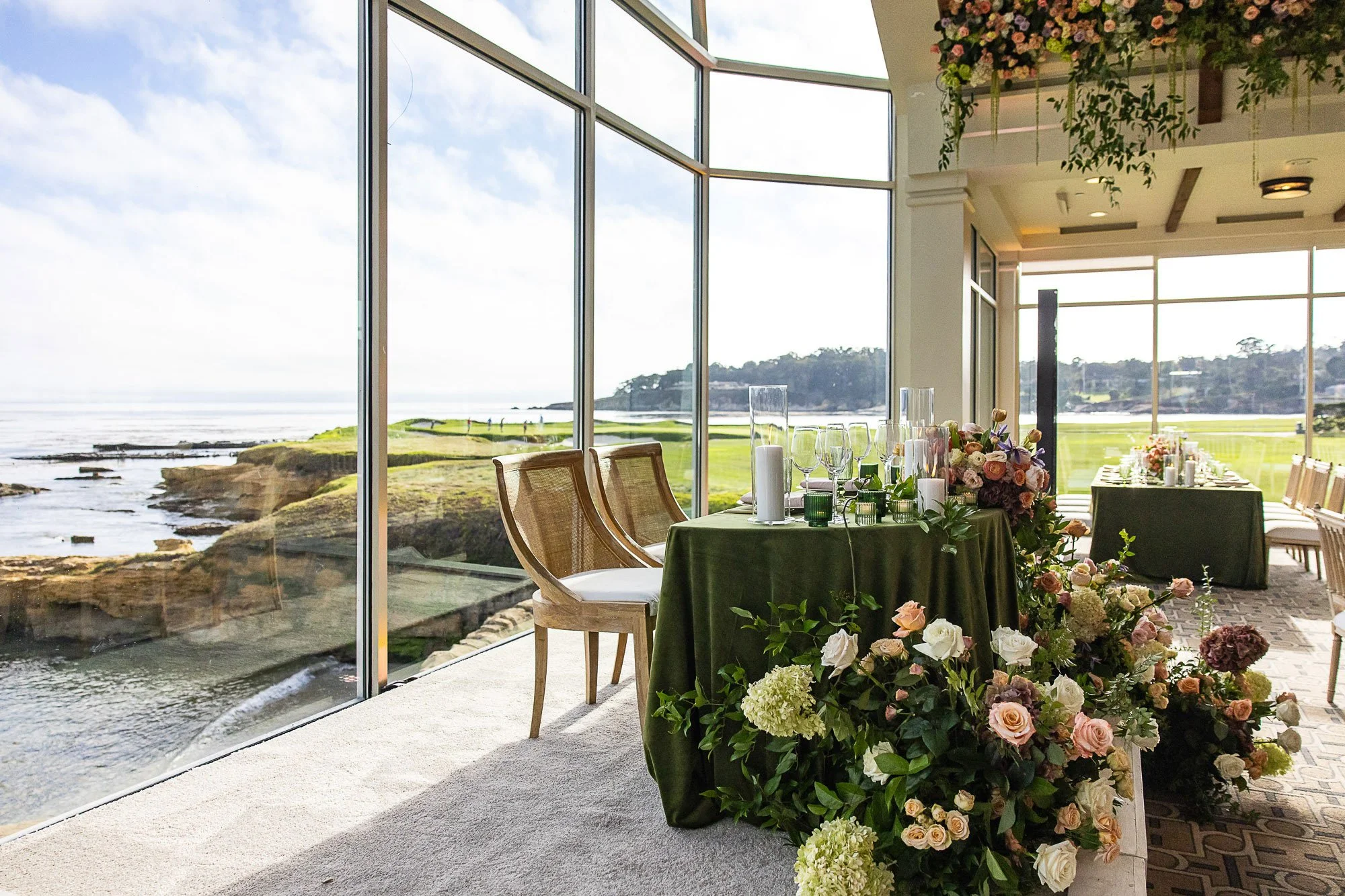 A banquet table decorated with flowers and candles inside a large glass-walled building overlooking a coastal landscape with grassy fields and a rocky shoreline at the Beach and Tennis Club in Pebble Beach, CA.