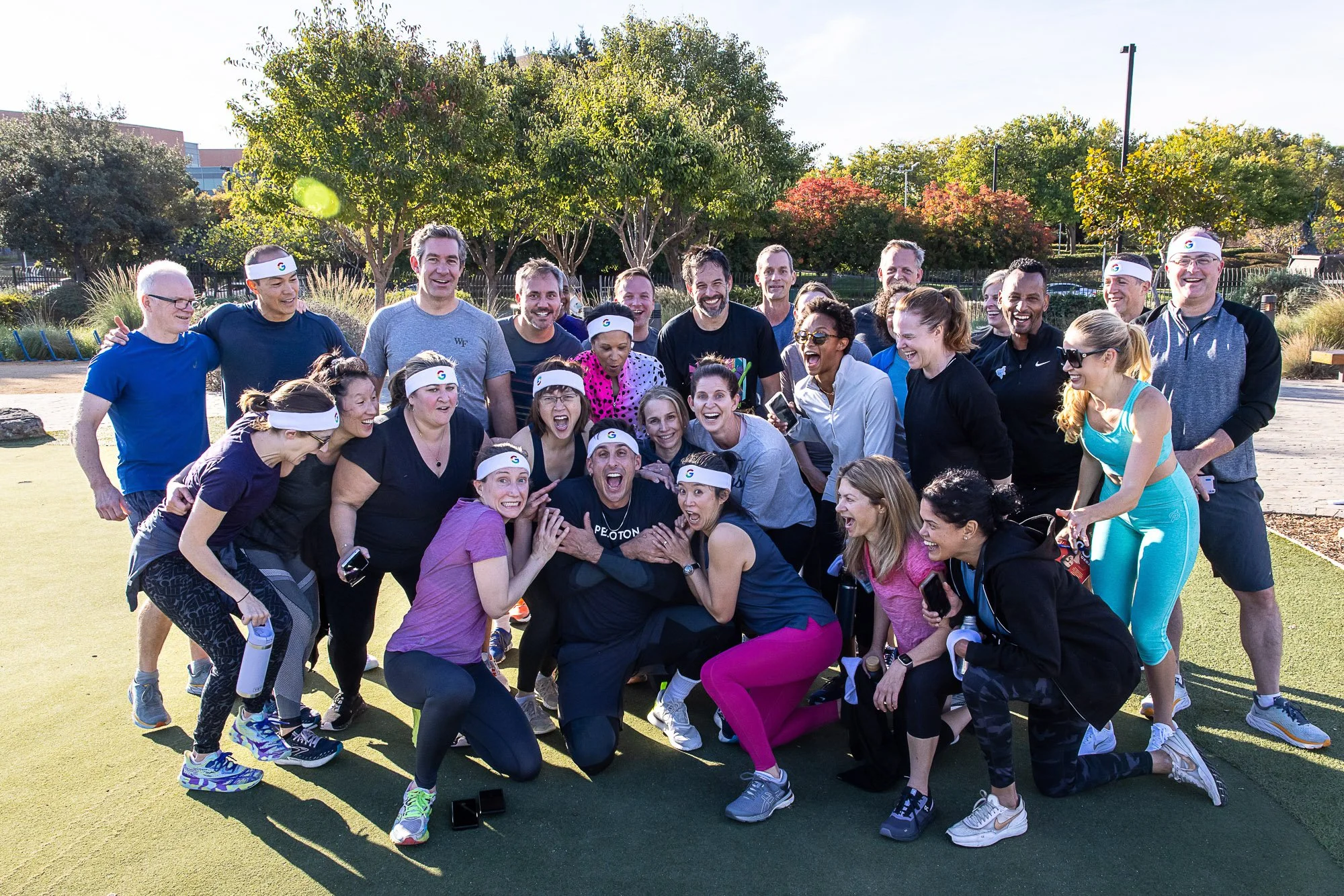A group of people gathered outdoors on a sunny day, smiling and laughing, dressed in athletic clothing, some wearing headbands with the Google logo, participating in a group fitness activity on a green field with trees and a fence in the background.