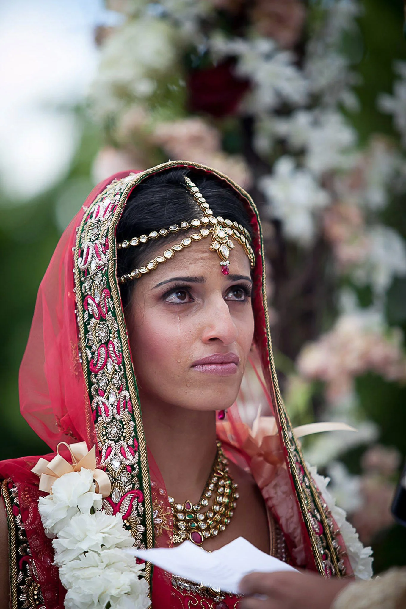 A woman dressed in traditional Indian wedding attire, including a red embroidered saree, jewelry, and a floral garland, is looking upwards during a ceremony outdoors with greenery in the background.