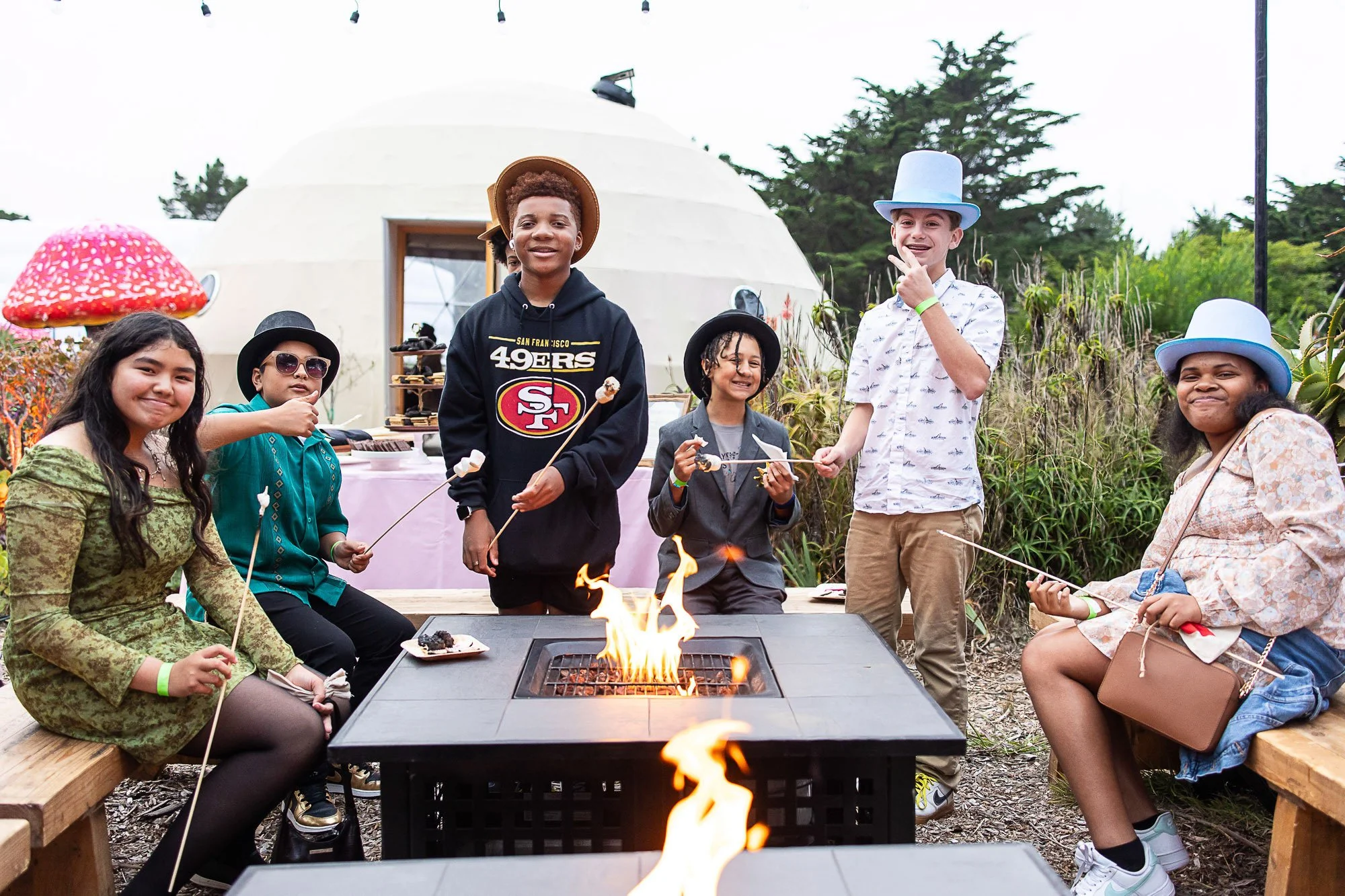 Group of kids sitting and standing around a fire pit at an outdoor gathering, some holding marshmallows on sticks, with a small round building and trees in the background.