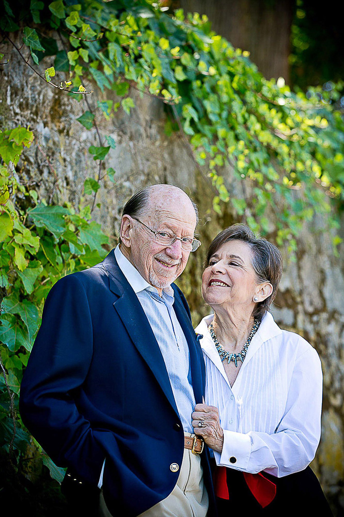 Older man and woman smiling and holding hands outdoors among green ivy and trees.