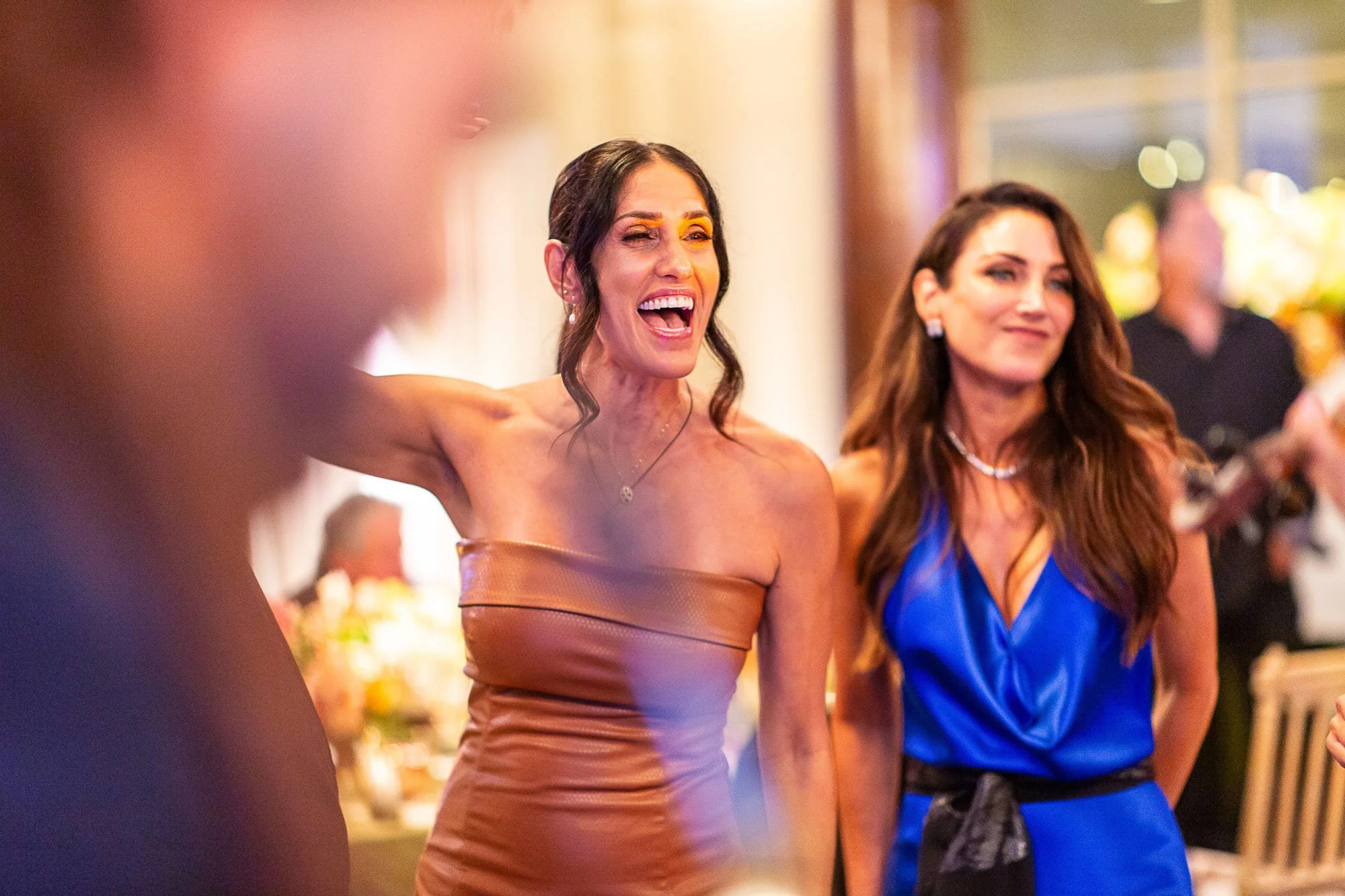 Two women at a social event, one wearing a strapless brown dress and the other in a blue dress, smiling and engaging with others at the Beach and Tennis Club in Pebble Beach, CA.