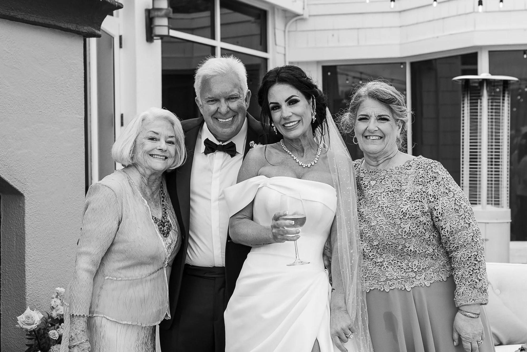 A group of five people at a wedding reception, including a bride in a strapless wedding gown and a groom in a tuxedo with a bowtie, standing together and smiling for a photo. The bride is holding a glass of champagne.