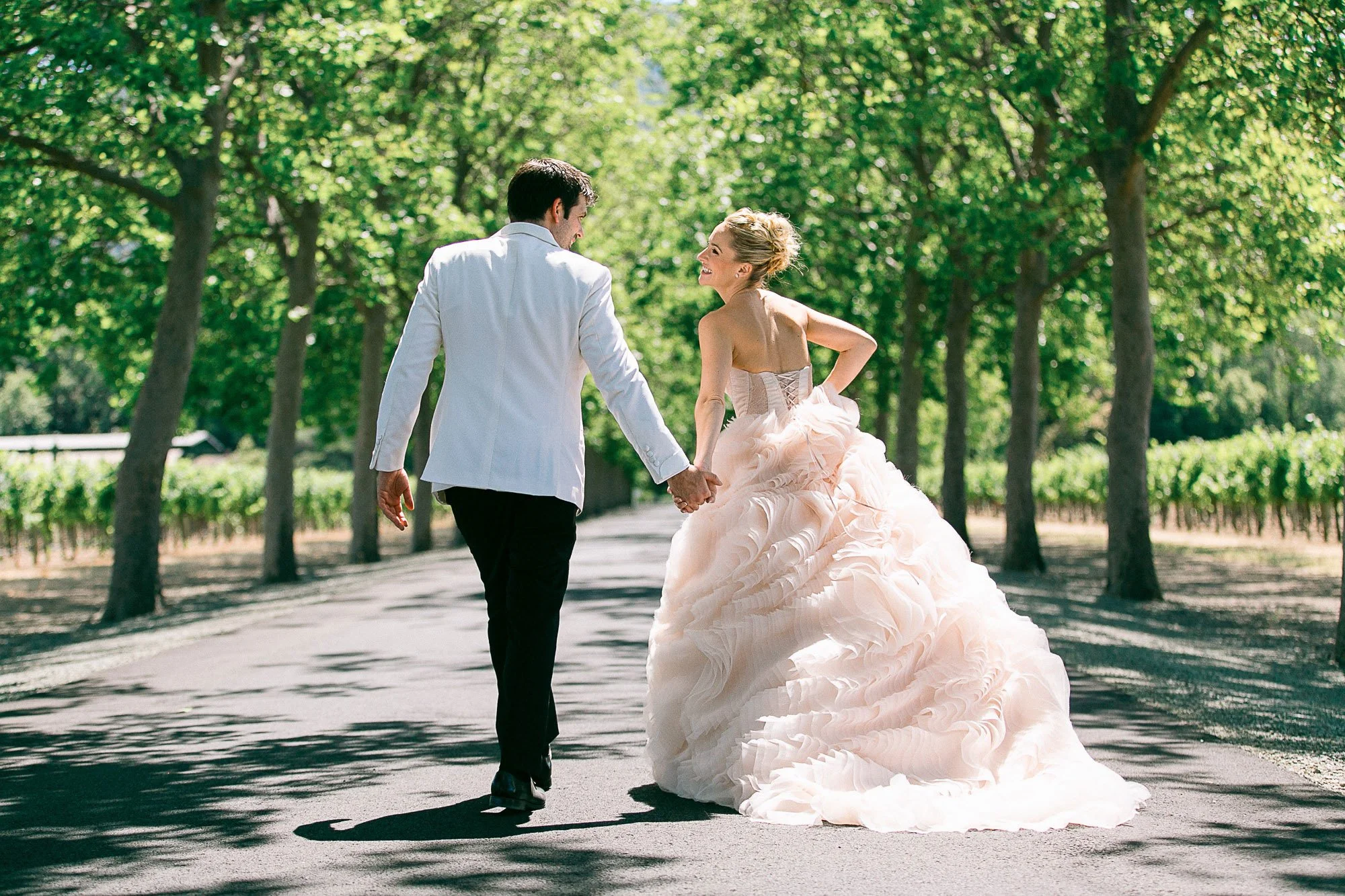 Bride and groom walking hand in hand down a tree-lined vineyard road in Napa Valley