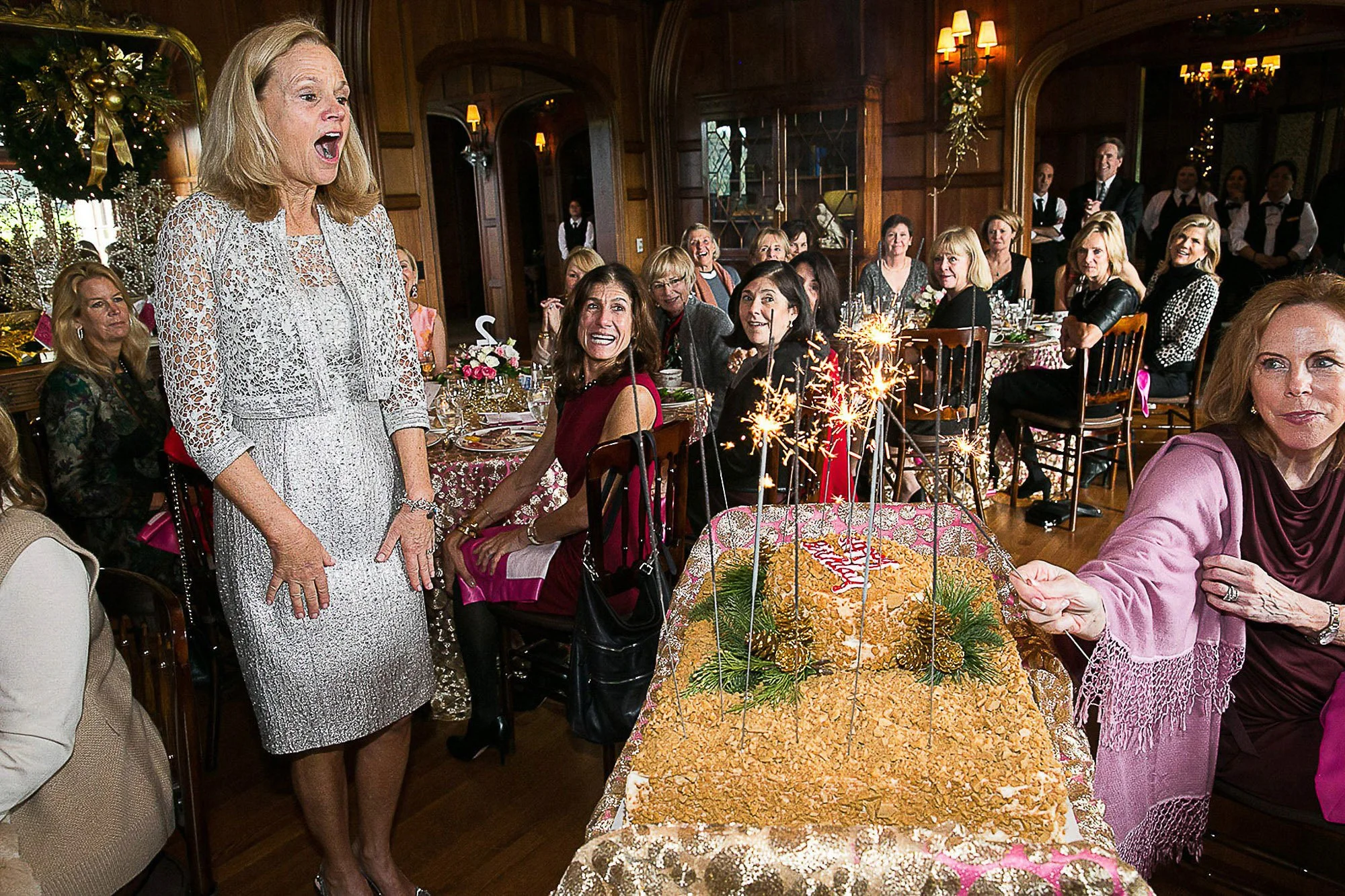 Women celebrating a birthday in a decorated, cozy, wooden-paneled room; one woman stands and sings, surrounded by friends at a table with a large birthday cake and sparklers.