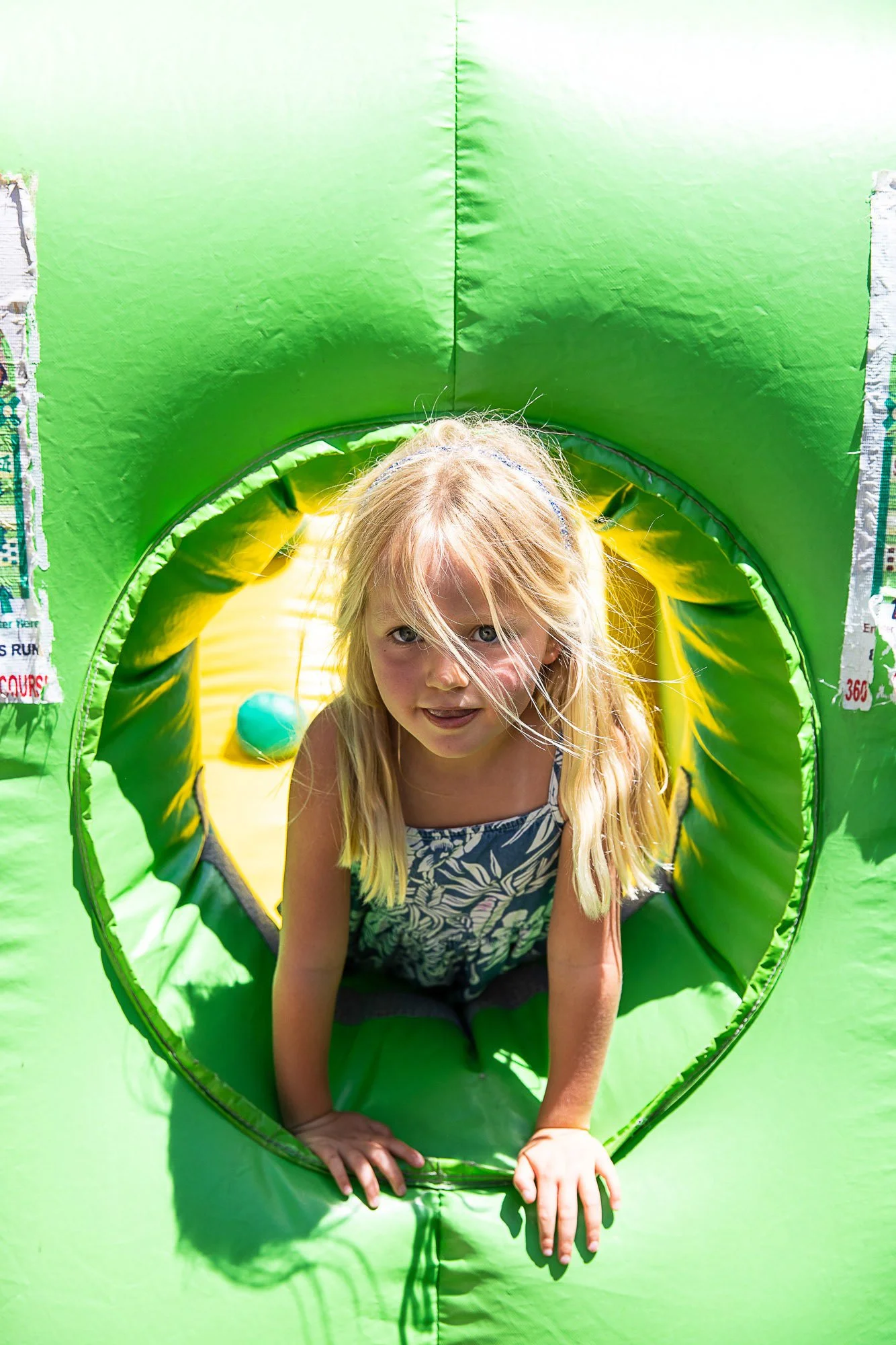 A girl with long blonde hair crawling through a green inflatable tunnel with yellow interior, outdoors on a sunny day.