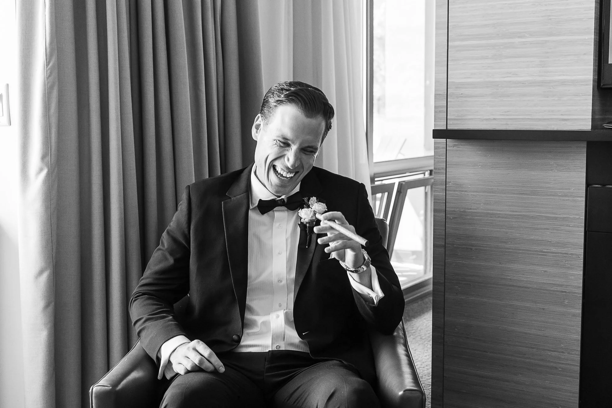 A groom dressed in a tuxedo laughing while adjusting his boutonniere, sitting in a modern room with curtains and window in the background at Cavallo Point in Sausalito, CA.