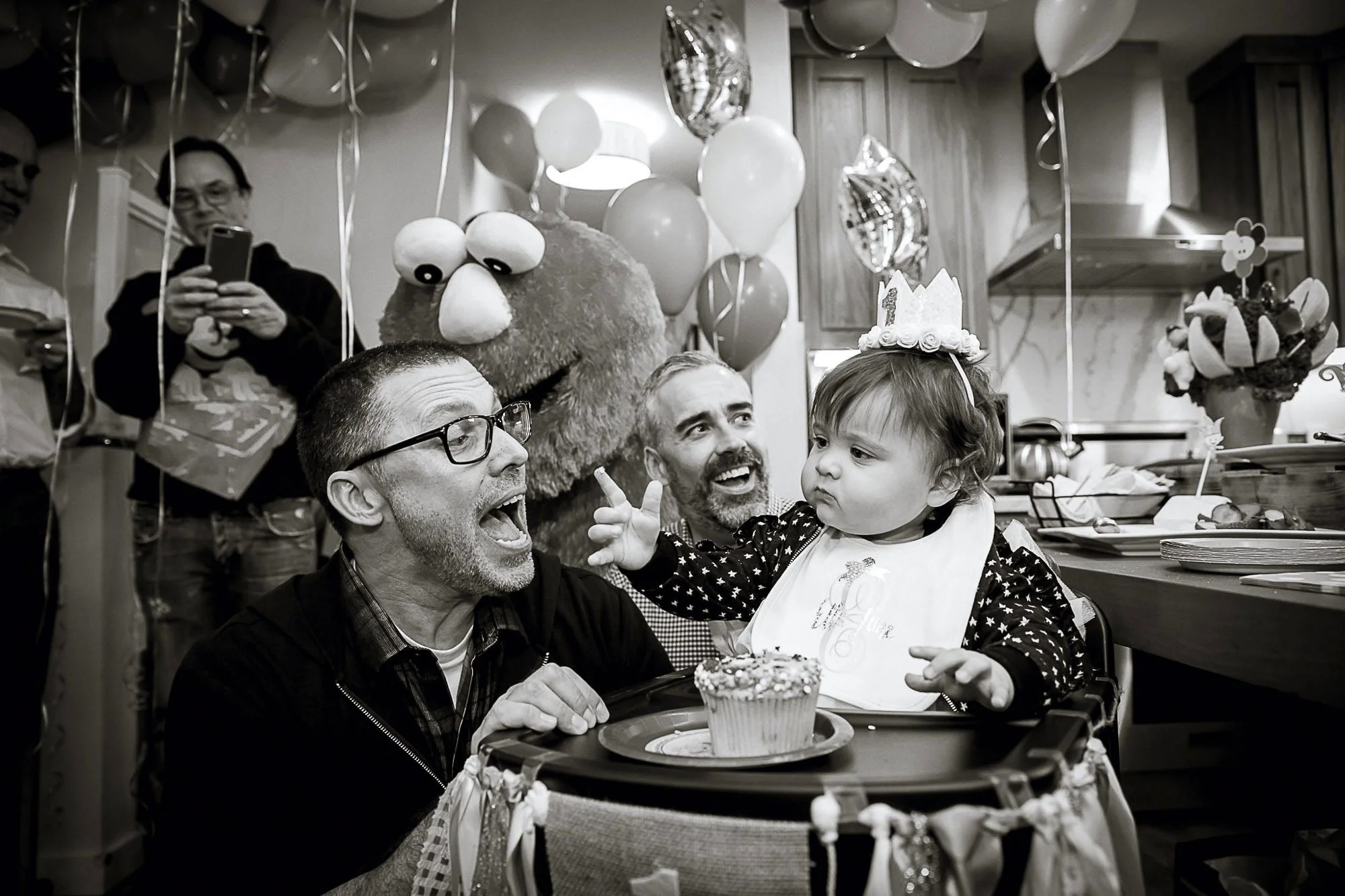 A birthday celebration with children and adults, including a young girl wearing a birthday crown. The girl is reaching towards a cupcake on a tray held by a man. There are balloons and decorations in the background, with some individuals taking photo