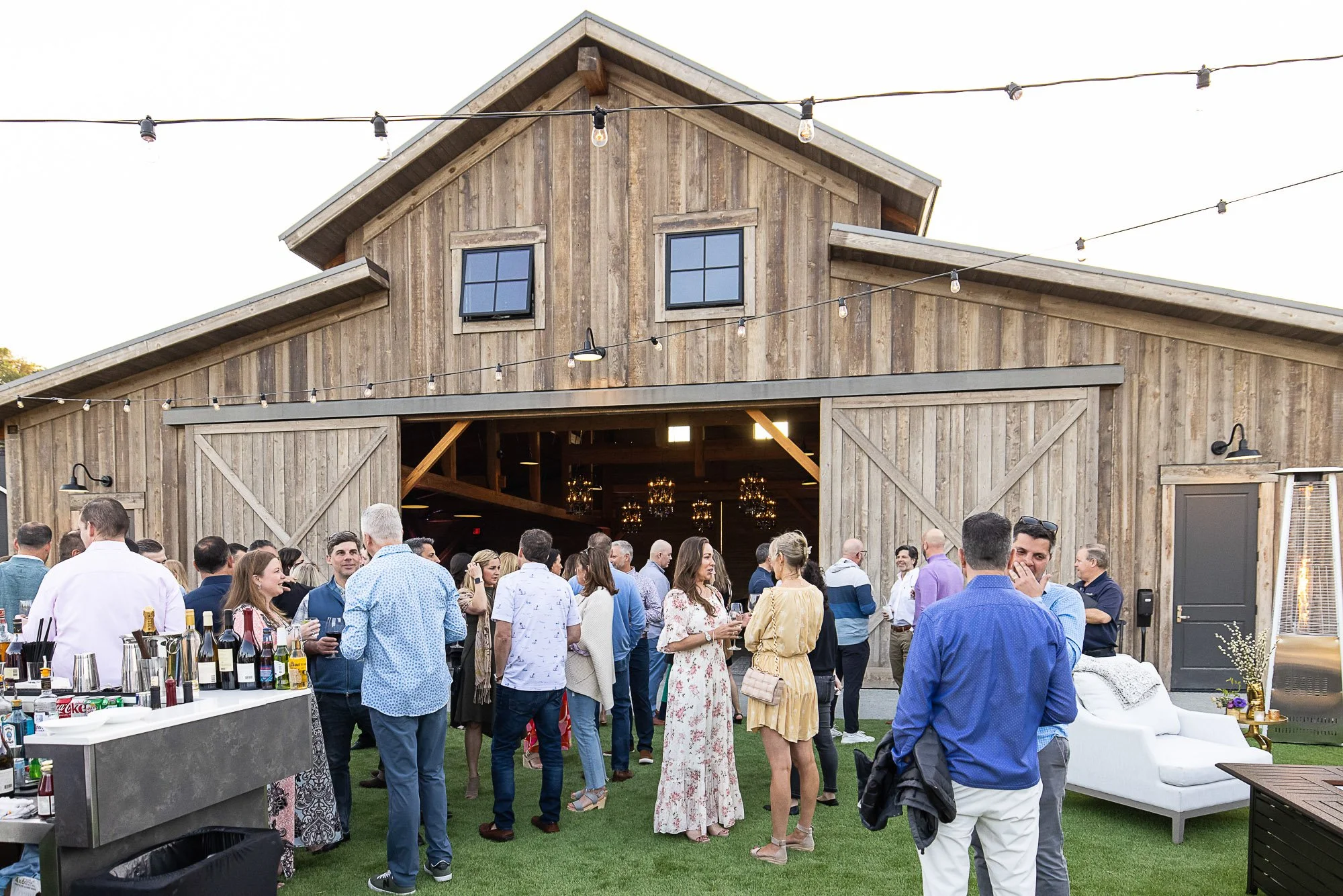 A gathering of people outside a rustic barn with string lights overhead, socializing at a party, with a table of wine bottles and drinks in the foreground at the Napa Valley Four Seasons Hotel.