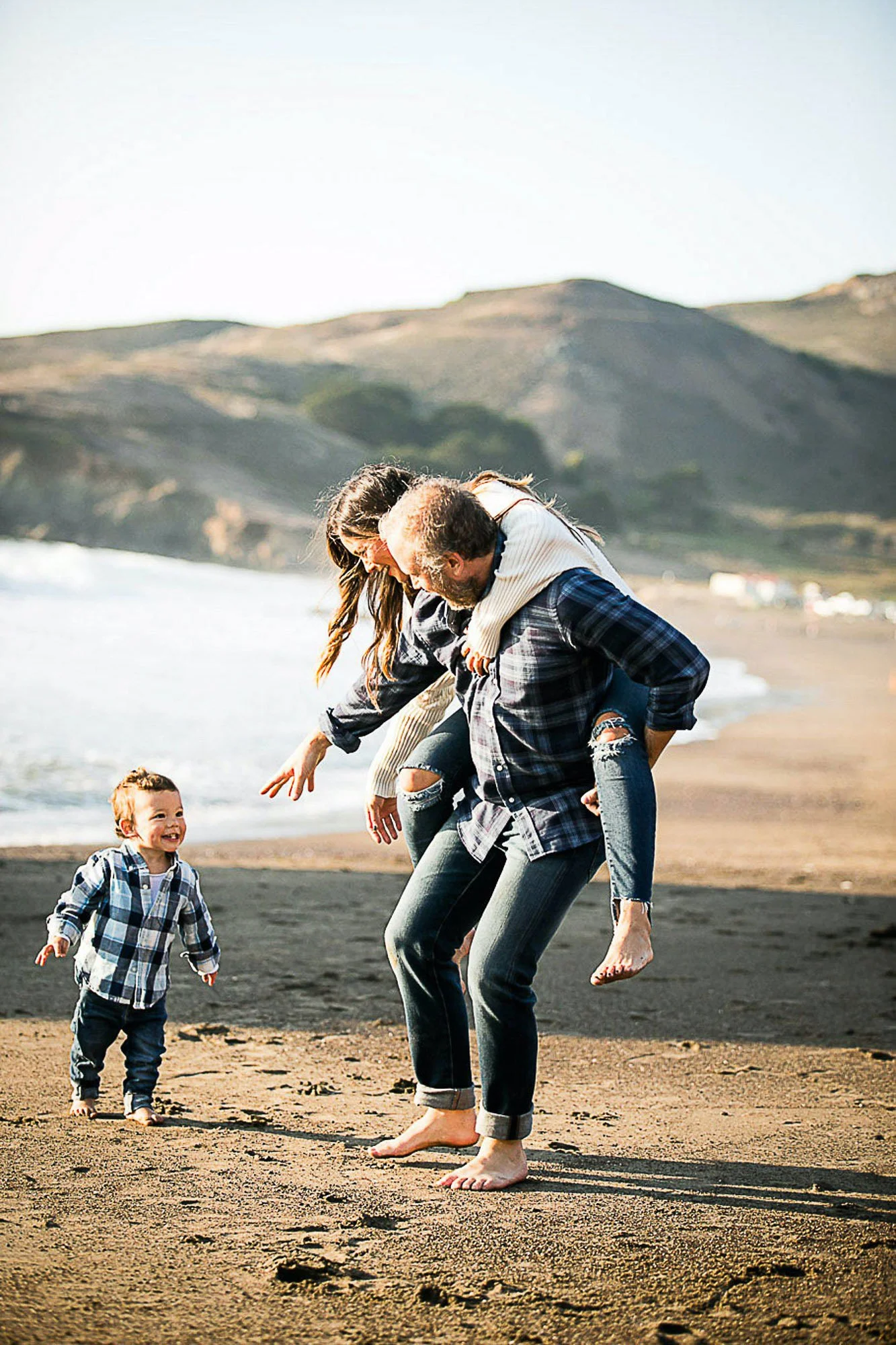 A family playing on a beach with mountains in the background. The man is giving a piggyback ride to a woman, while a young boy in a plaid shirt runs beside them.