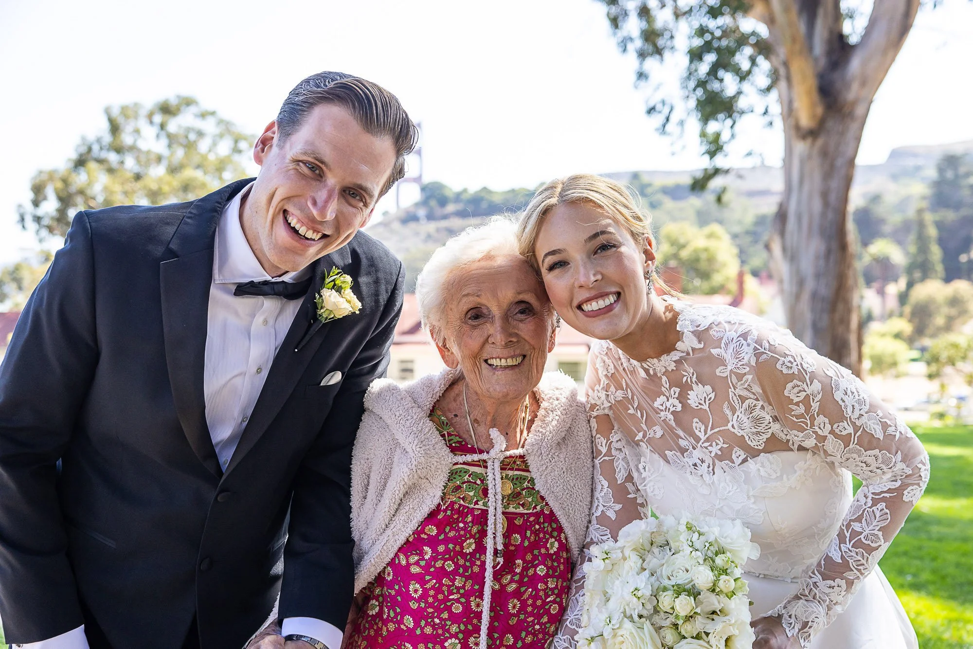Group of four smiling women, including a bride in a wedding dress, posing outdoors with trees and a hillside in the background at Cavallo Point in Sausalito, CA.