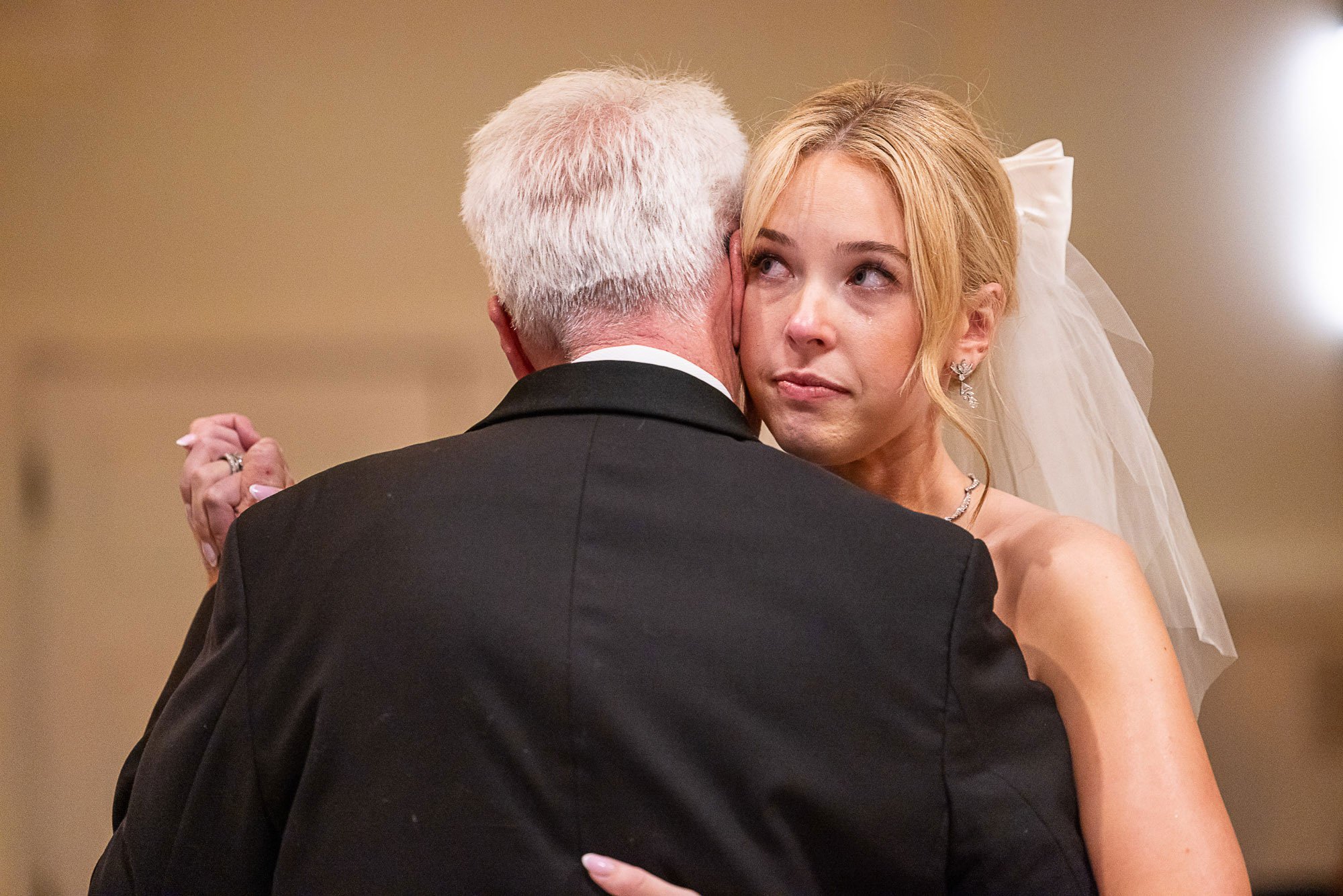 A bride and an older man, possibly her father, sharing a dance at a wedding. The bride has blonde hair, wearing earrings, and a white dress with a veil. The older man has white hair and is dressed in a black suit.