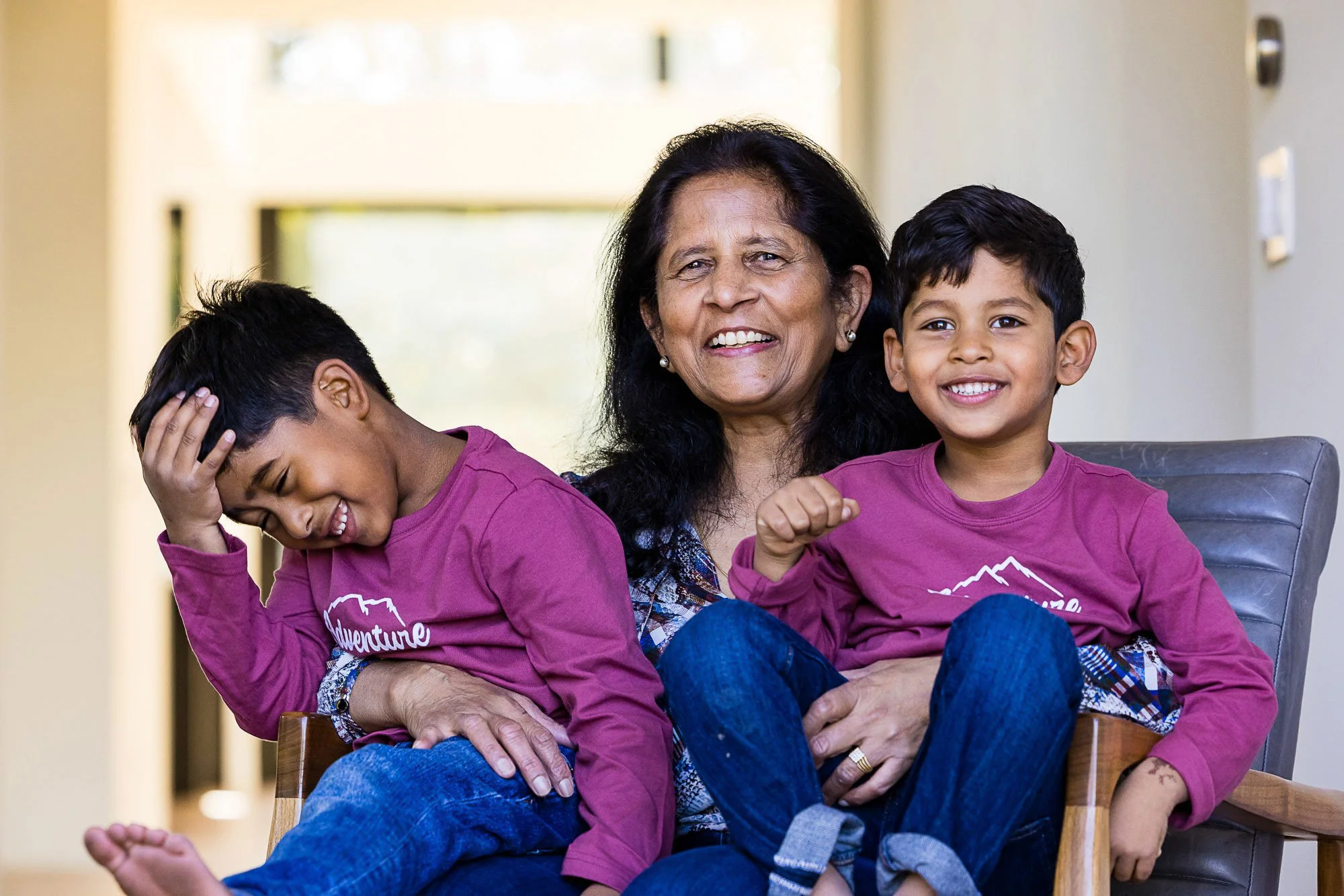 An elderly woman with black hair and earrings sitting on a chair with two young boys, one laughing and the other holding his head, all wearing matching purple shirts.