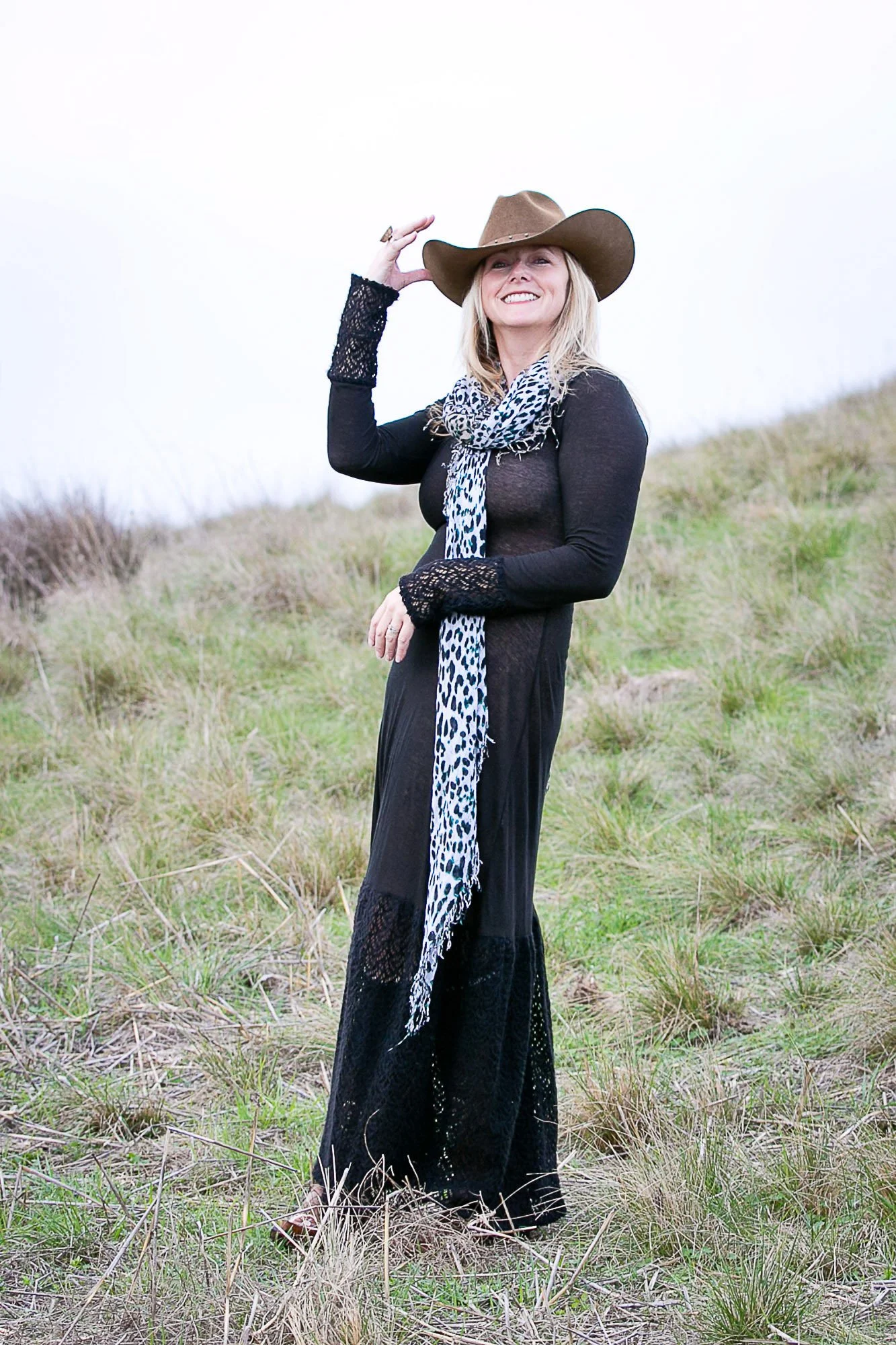 Tara Arrowood standing outdoors in a grassy field, wearing a large brown cowboy hat, a dark long-sleeved dress with lace details, and a scarf with a black and white leopard print pattern, smiling and holding her hat with one hand.