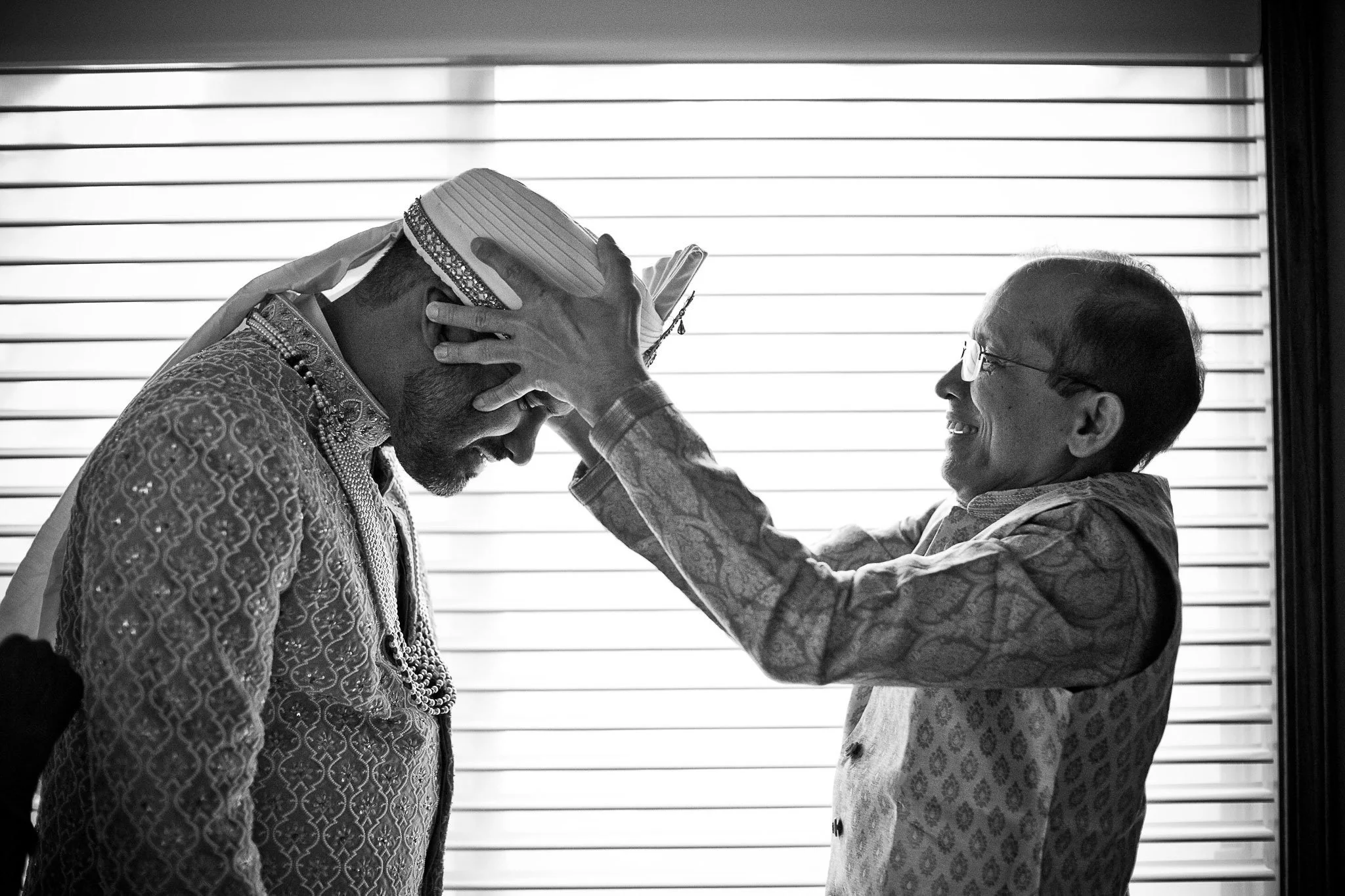 A black and white photo of a traditional Indian wedding ceremony with an elderly woman placing a head covering on a groom dressed in ornate attire.