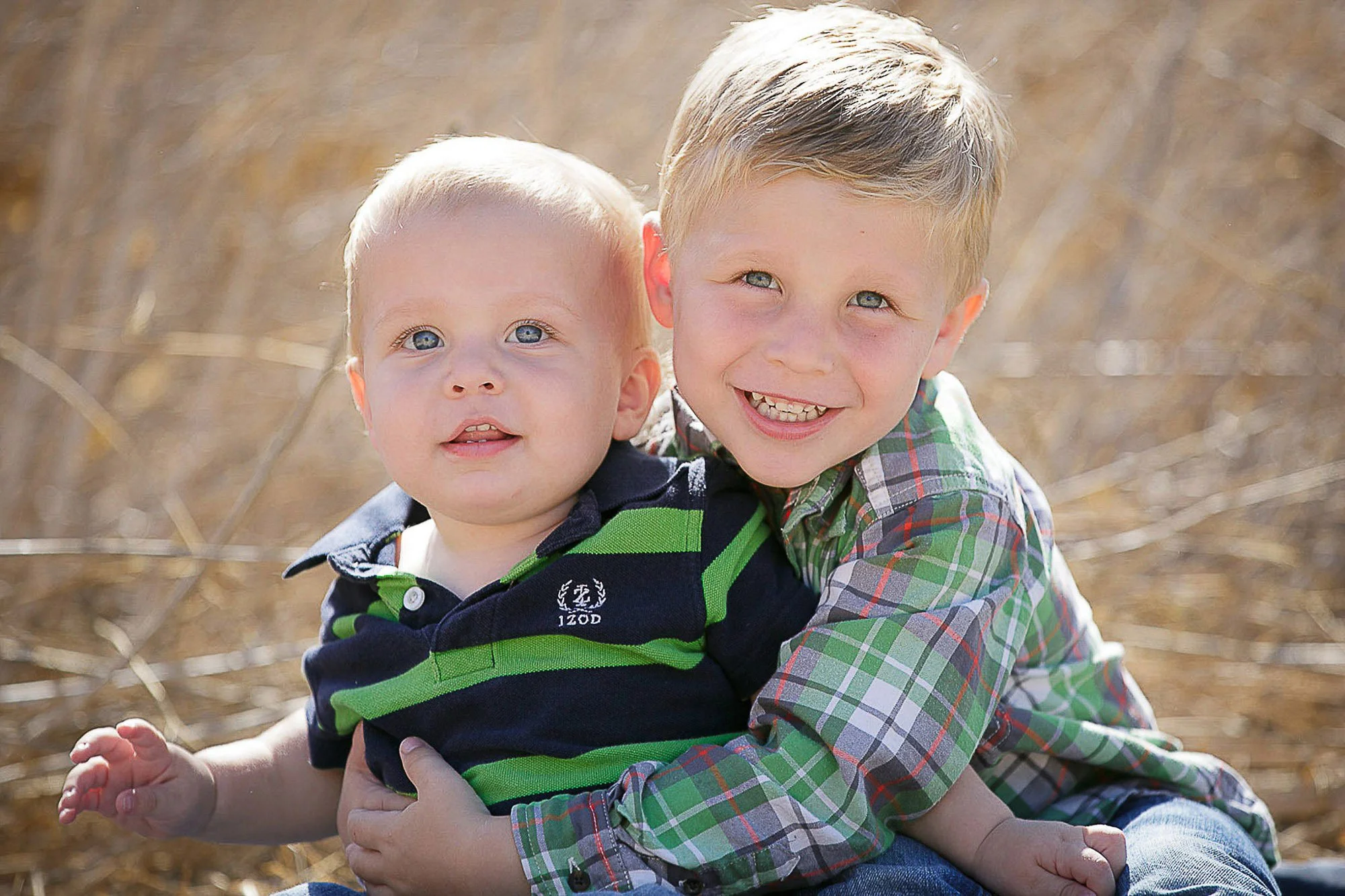 Two young boys smiling outdoors, one is older with blond hair and the other is younger with light blond hair, sitting and playing in a dry, grassy field.