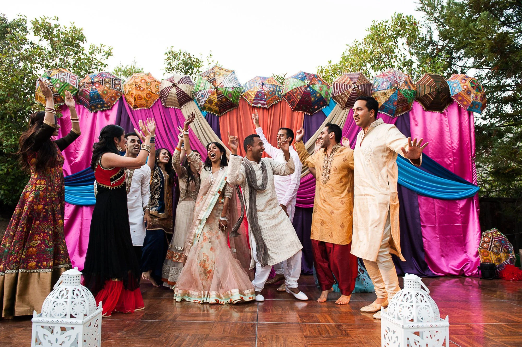People dancing and celebrating at an outdoor Indian wedding reception with colorful drapery and umbrellas in the background.