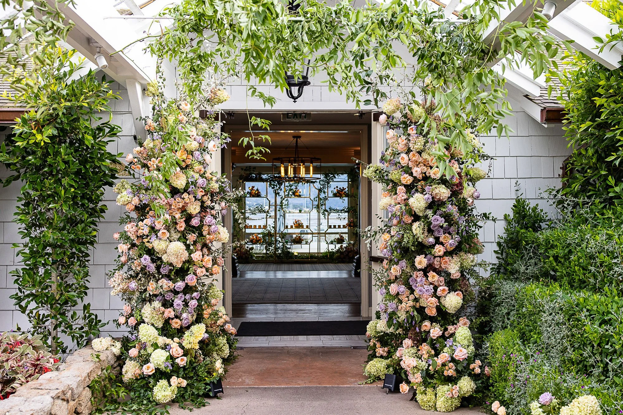 Archway decorated with cream, pink, and purple flowers leading into an indoor space with a view of hanging light fixtures and large windows at the Beach and Tennis Club in Pebble Beach, CA.