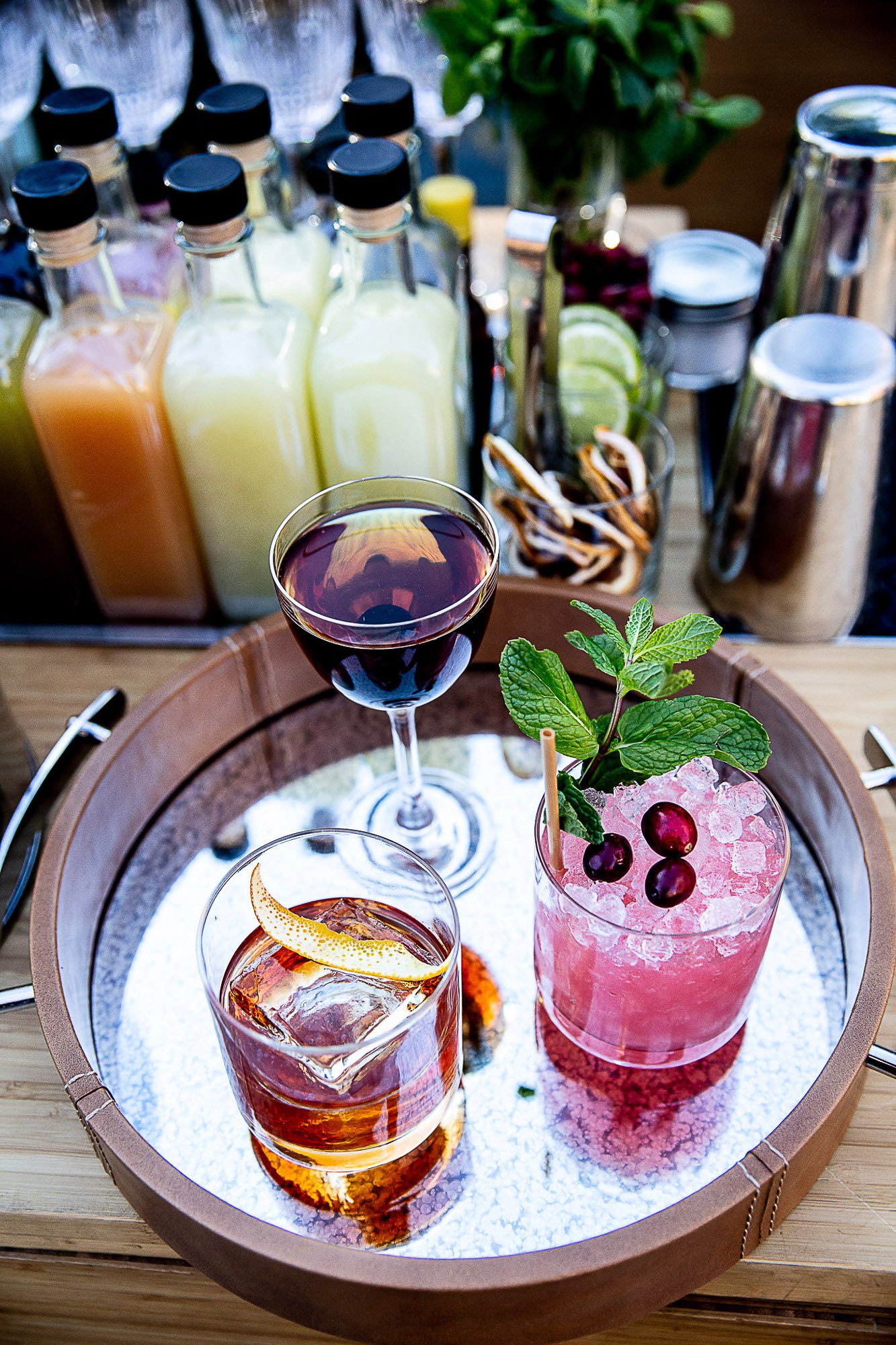 A tray with three colorful drinks: one garnished with berries and mint, another with orange peel and ice, and a glass of red wine, with bottled juices, lime slices, and a plant in the background.