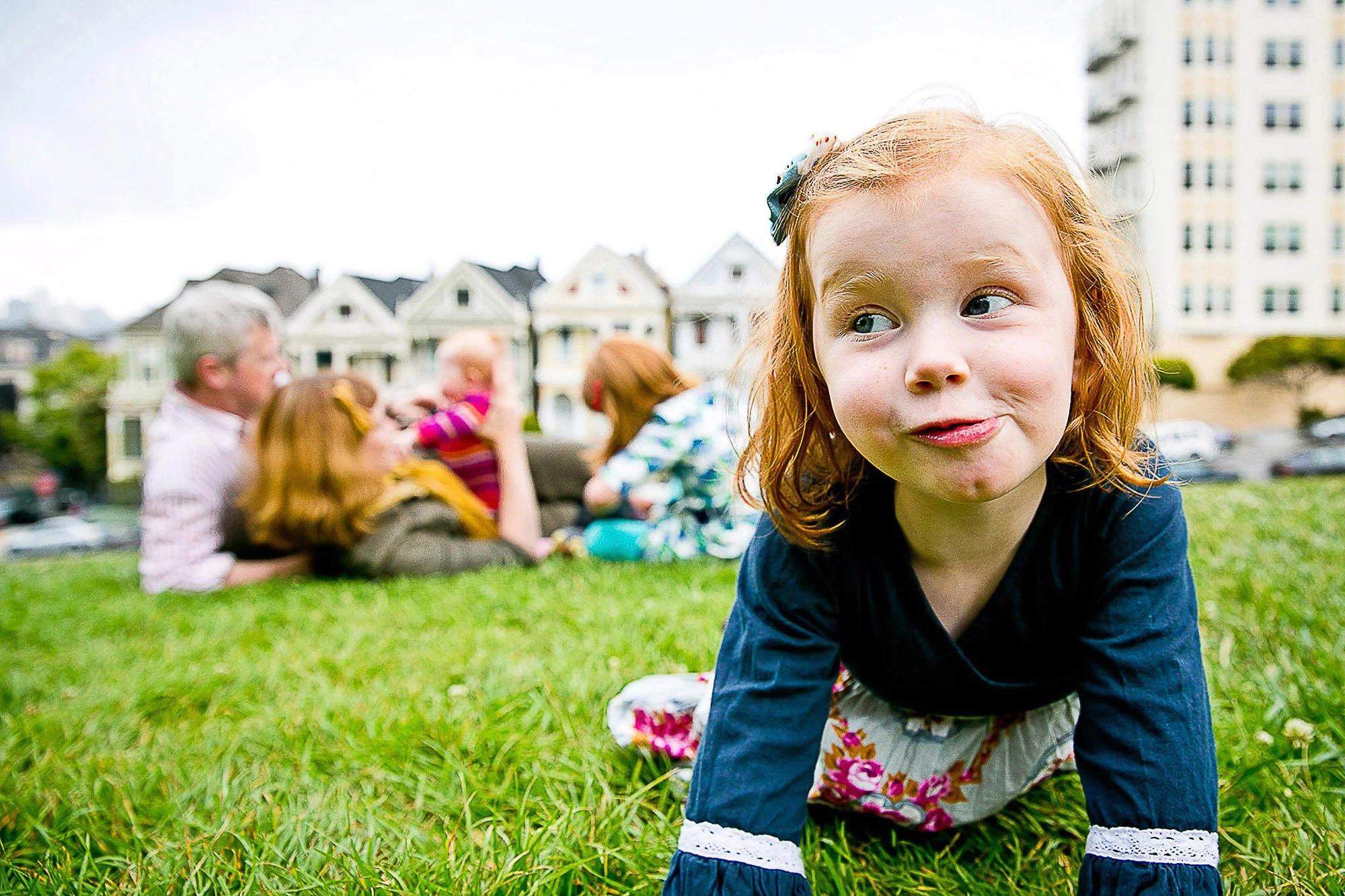 Young girl with red hair, wearing a black shirt and floral skirt, crawling on a grassy field with a group of adults and children in the background in Alamo Square in San Francisco and the painted ladies visible behind them.