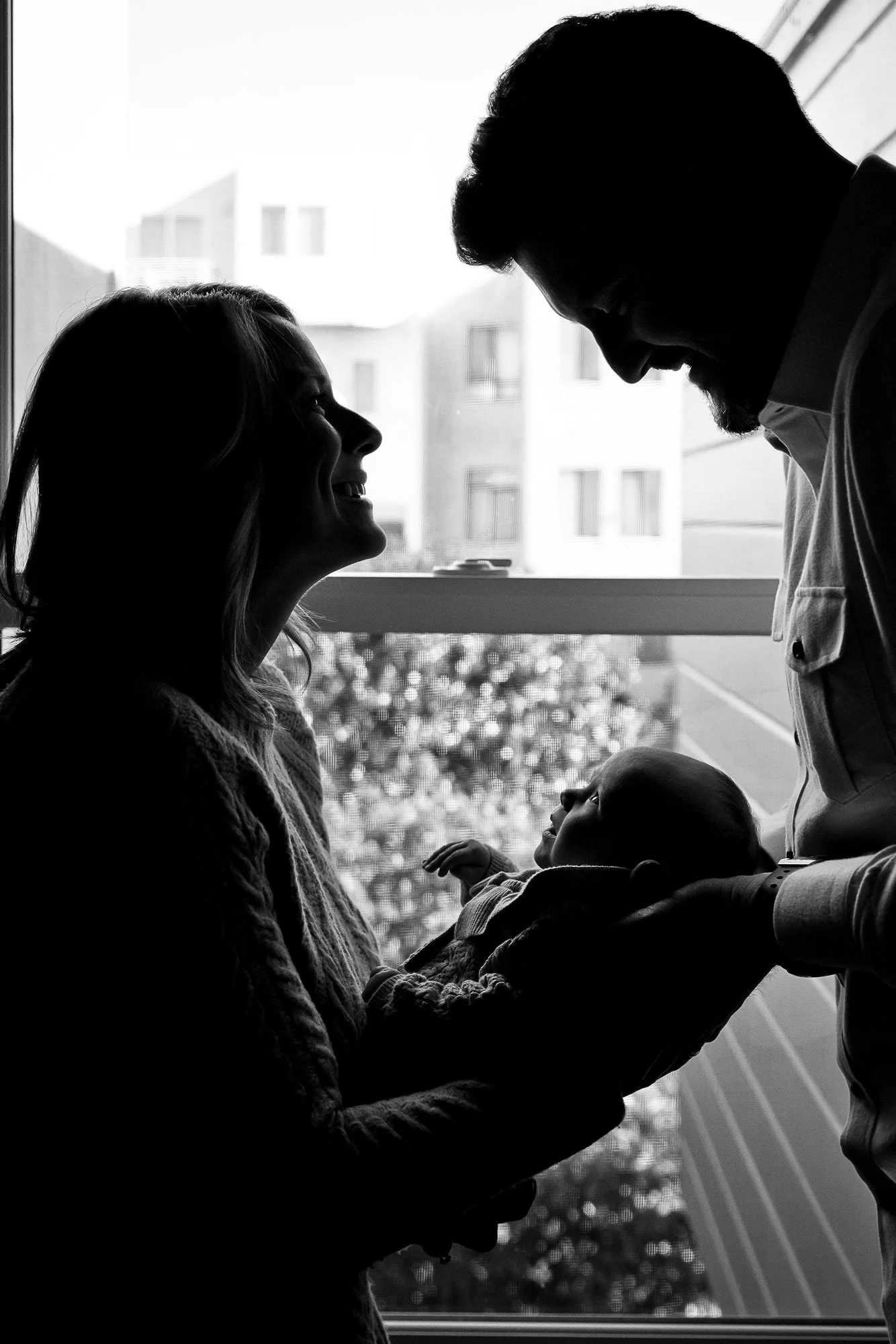 A black and white silhouette of a family with a woman, man, and baby, looking at each other in front of a window with buildings outside.